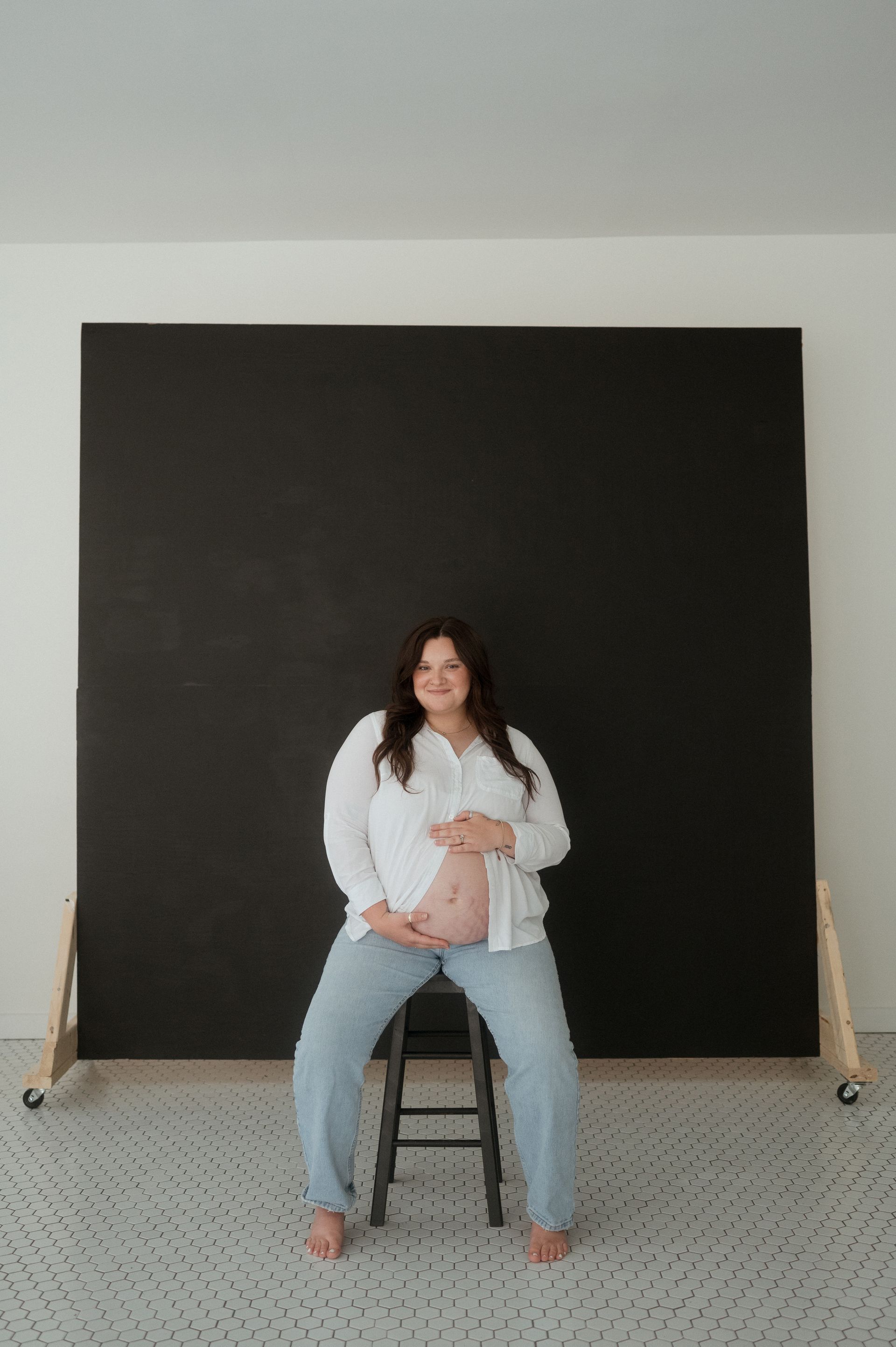 A pregnant woman is sitting on a stool in front of a blackboard.