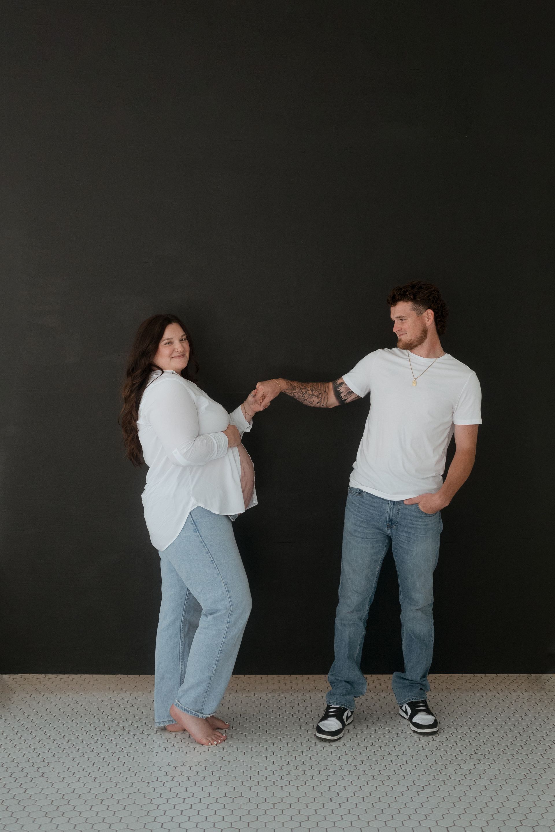 A man and a pregnant woman are dancing in front of a black wall.