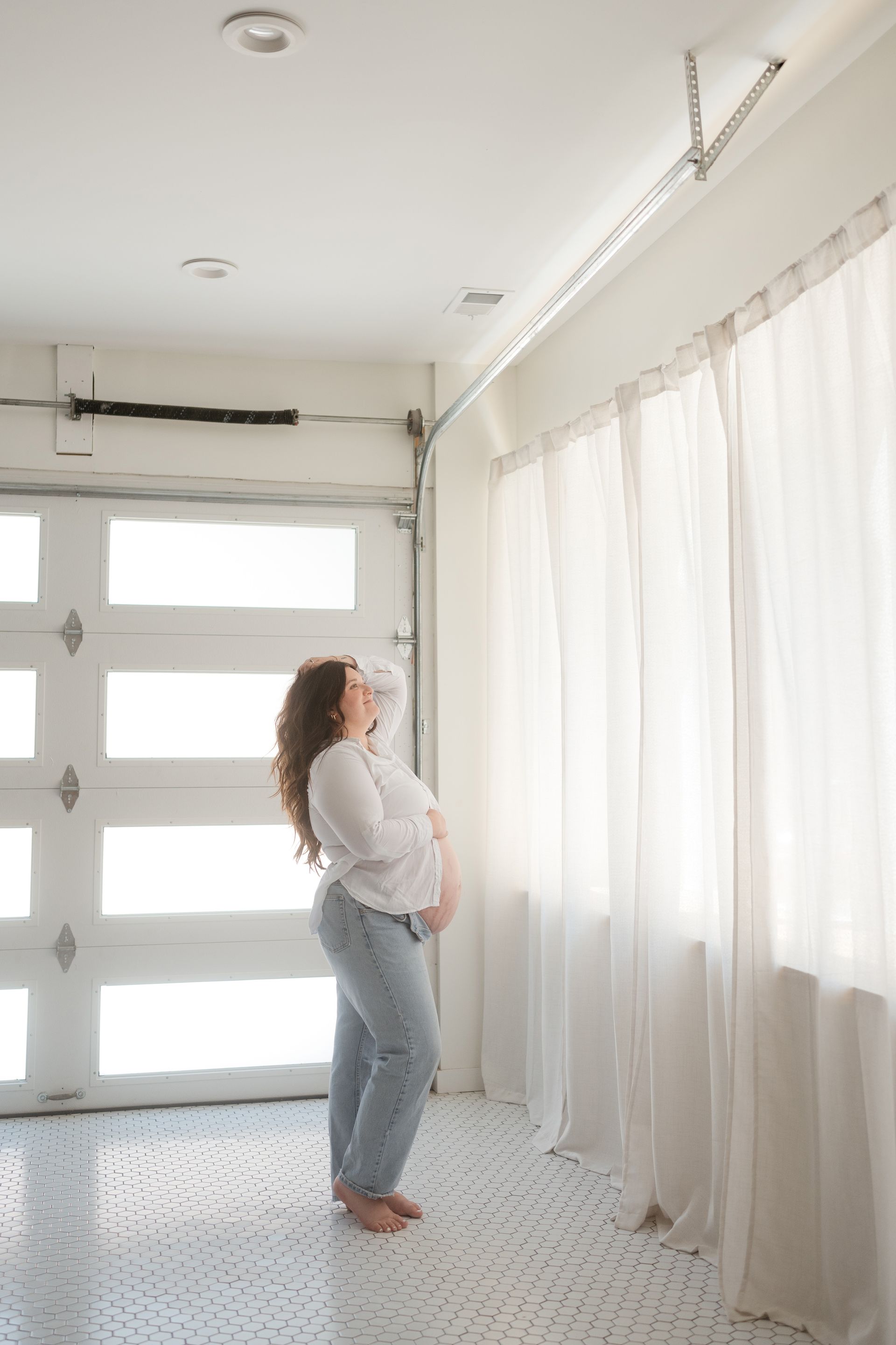 A pregnant woman is standing in a garage looking out the window.