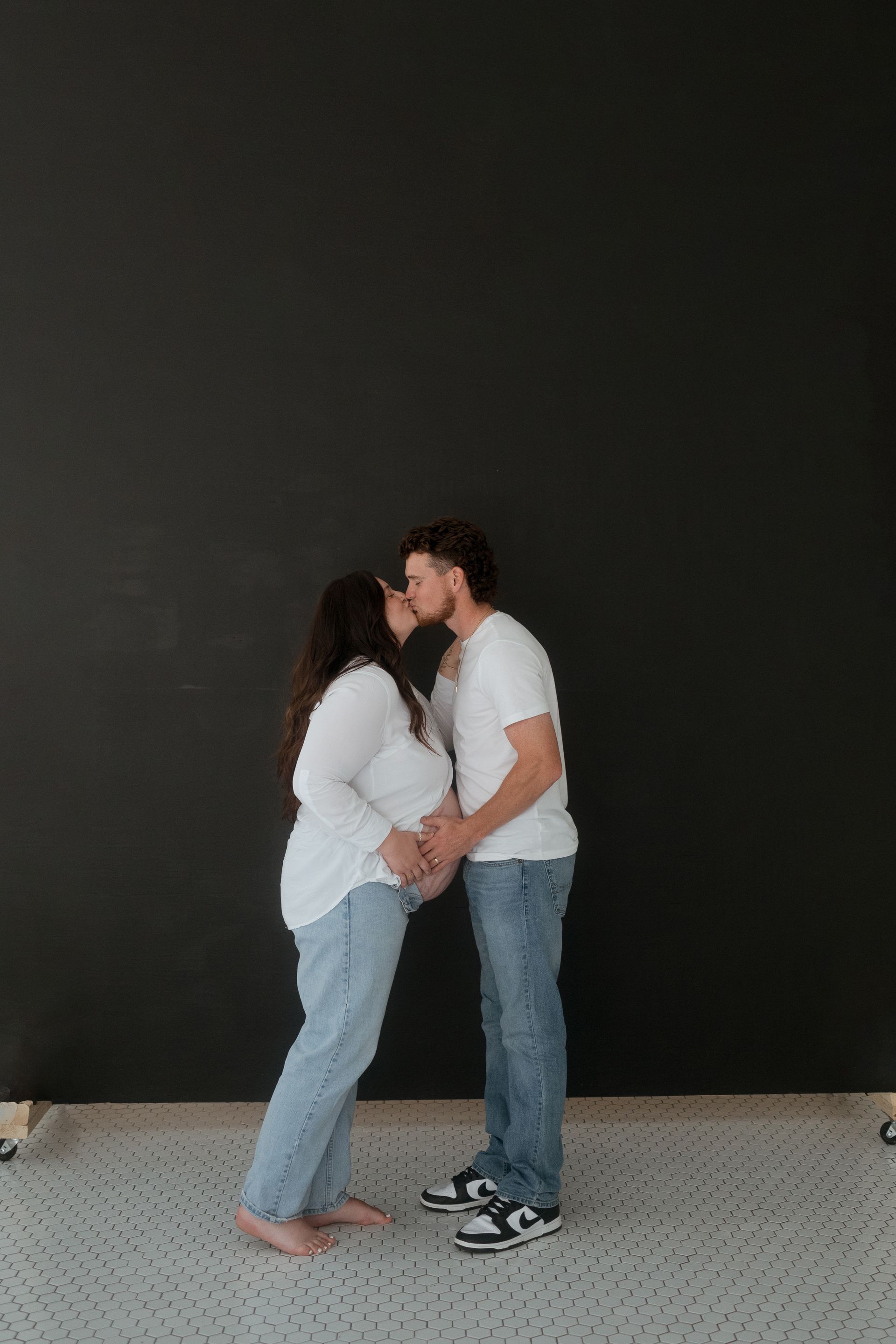 A man and a pregnant woman are kissing in front of a black wall.