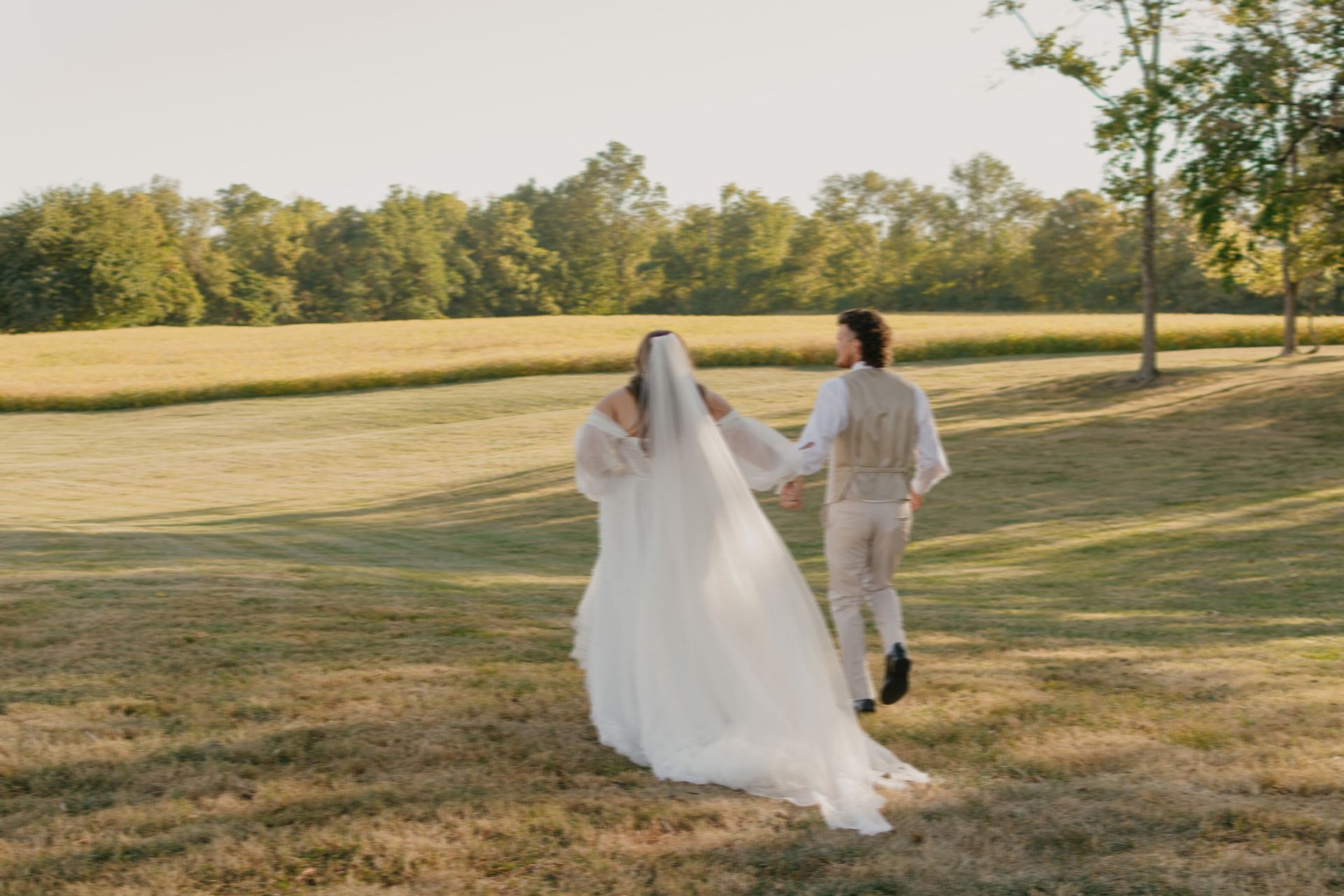 A bride and groom are walking through a field holding hands.