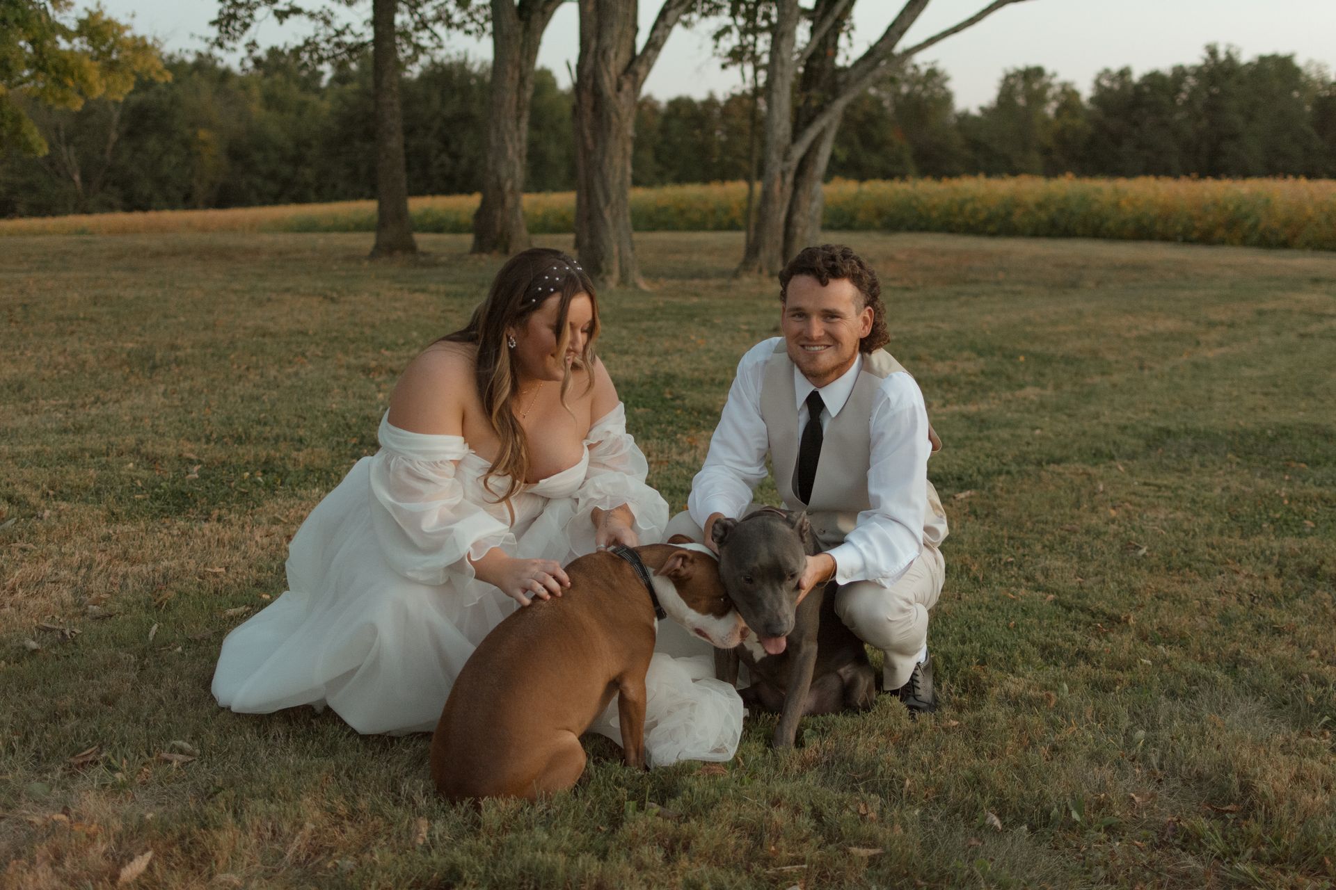 A bride and groom are kneeling in the grass with their dog.