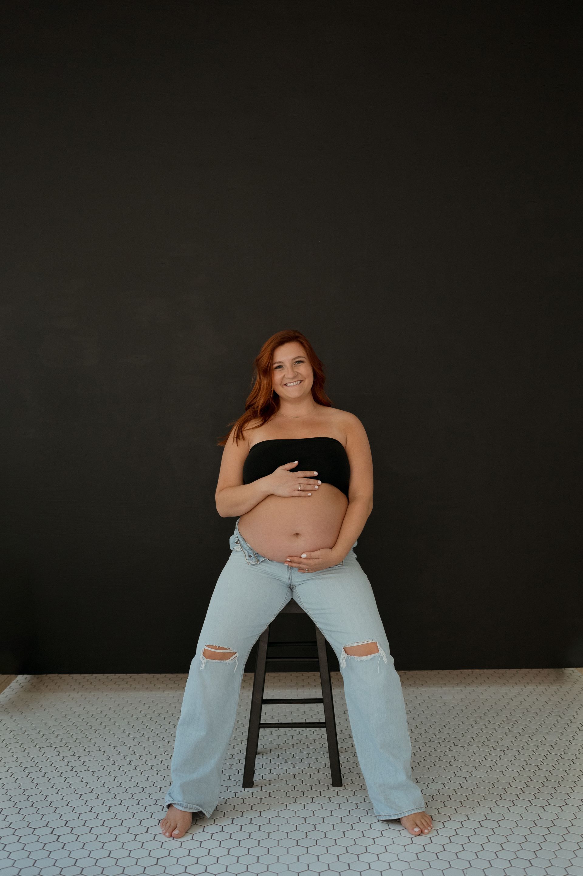 A pregnant woman is sitting on a stool holding her belly.