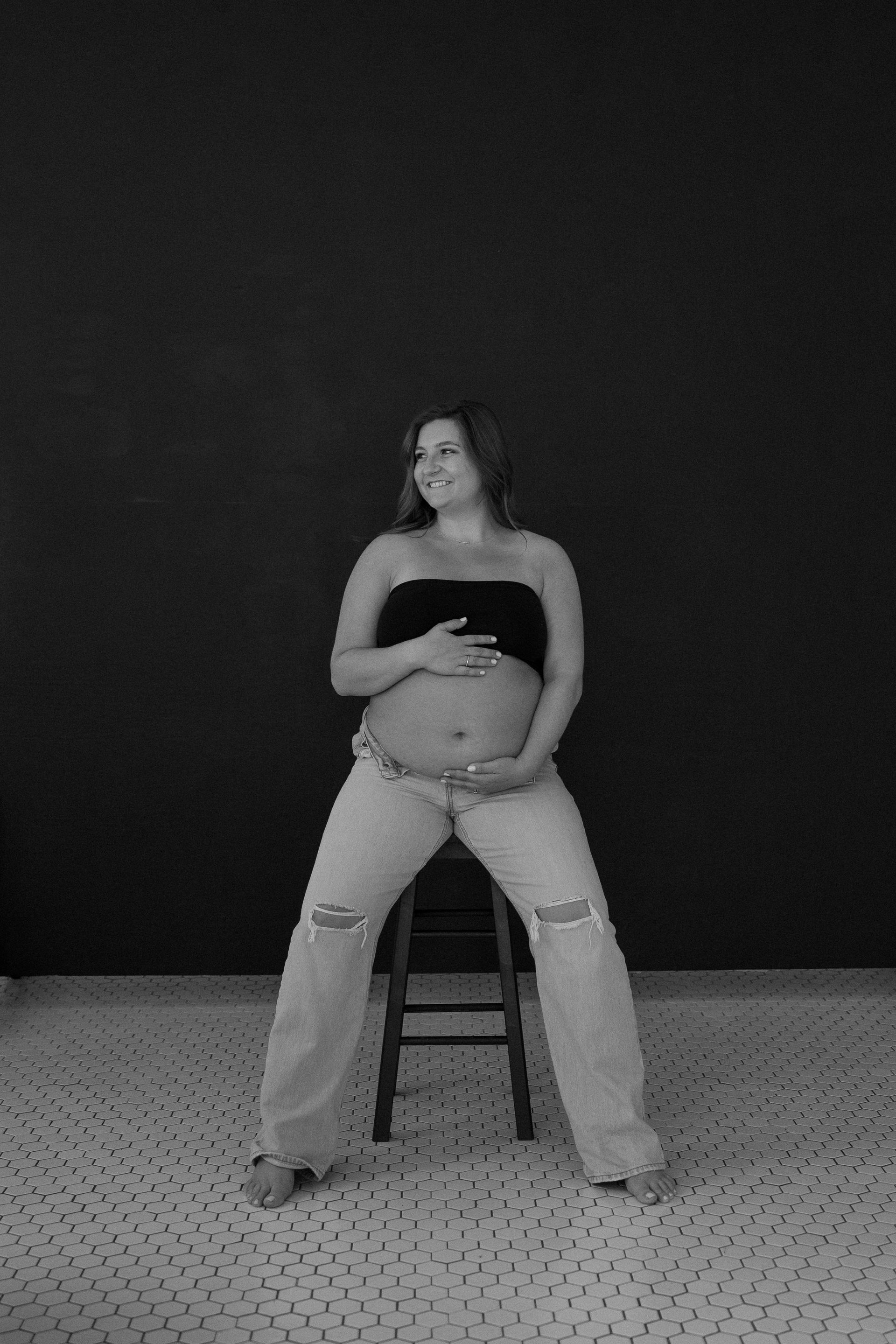 A pregnant woman is sitting on a stool in a black and white photo.