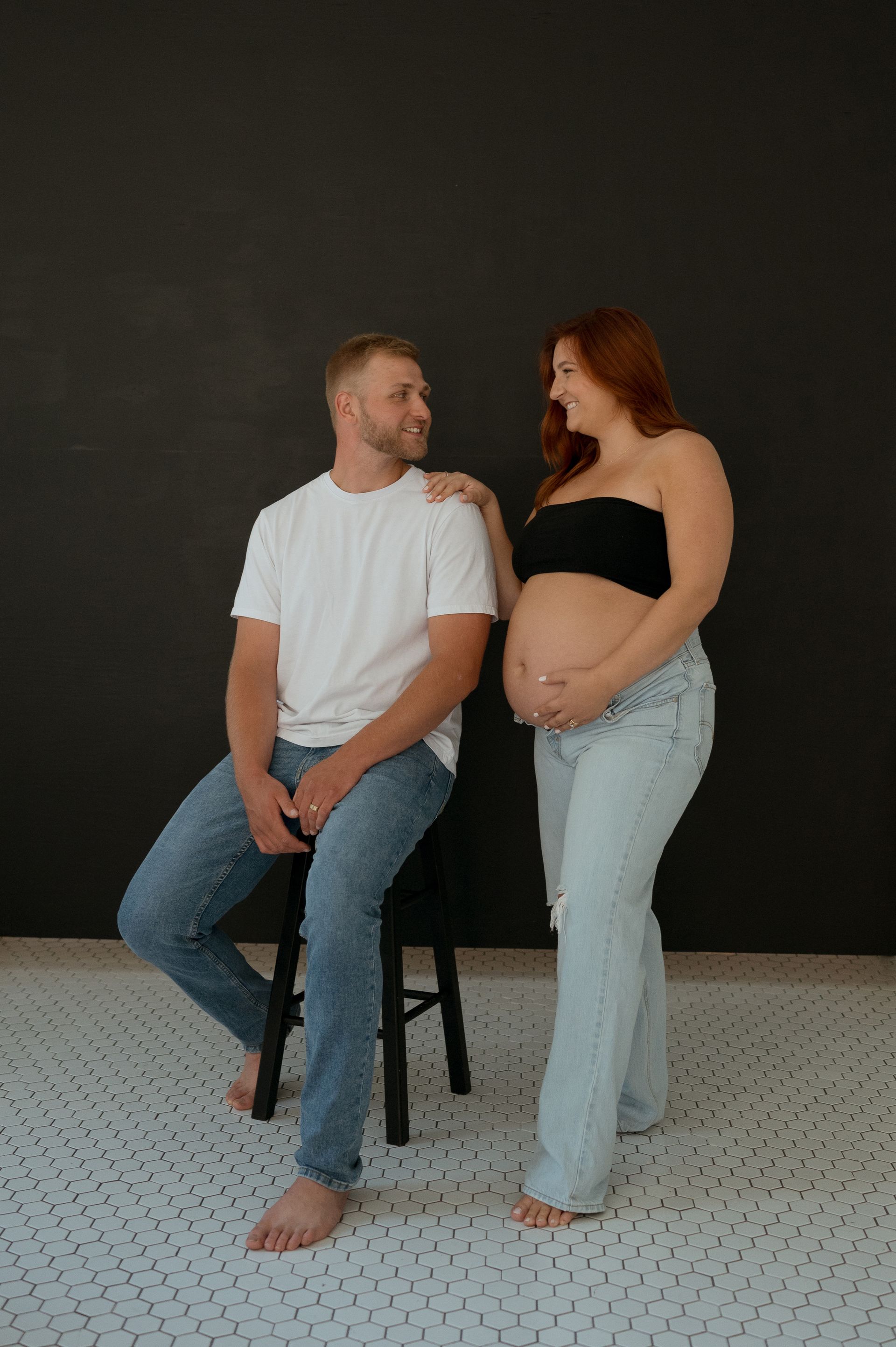 A man is sitting on a stool next to a pregnant woman.