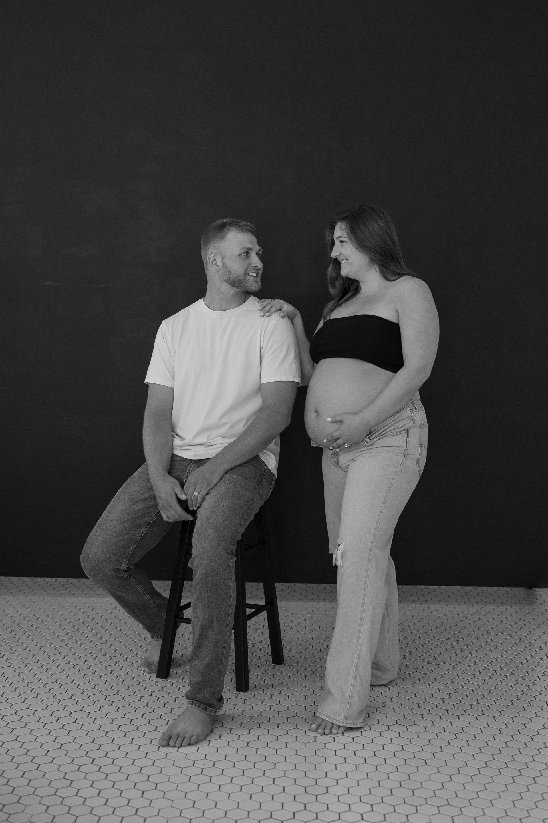 A man is sitting on a stool next to a pregnant woman.