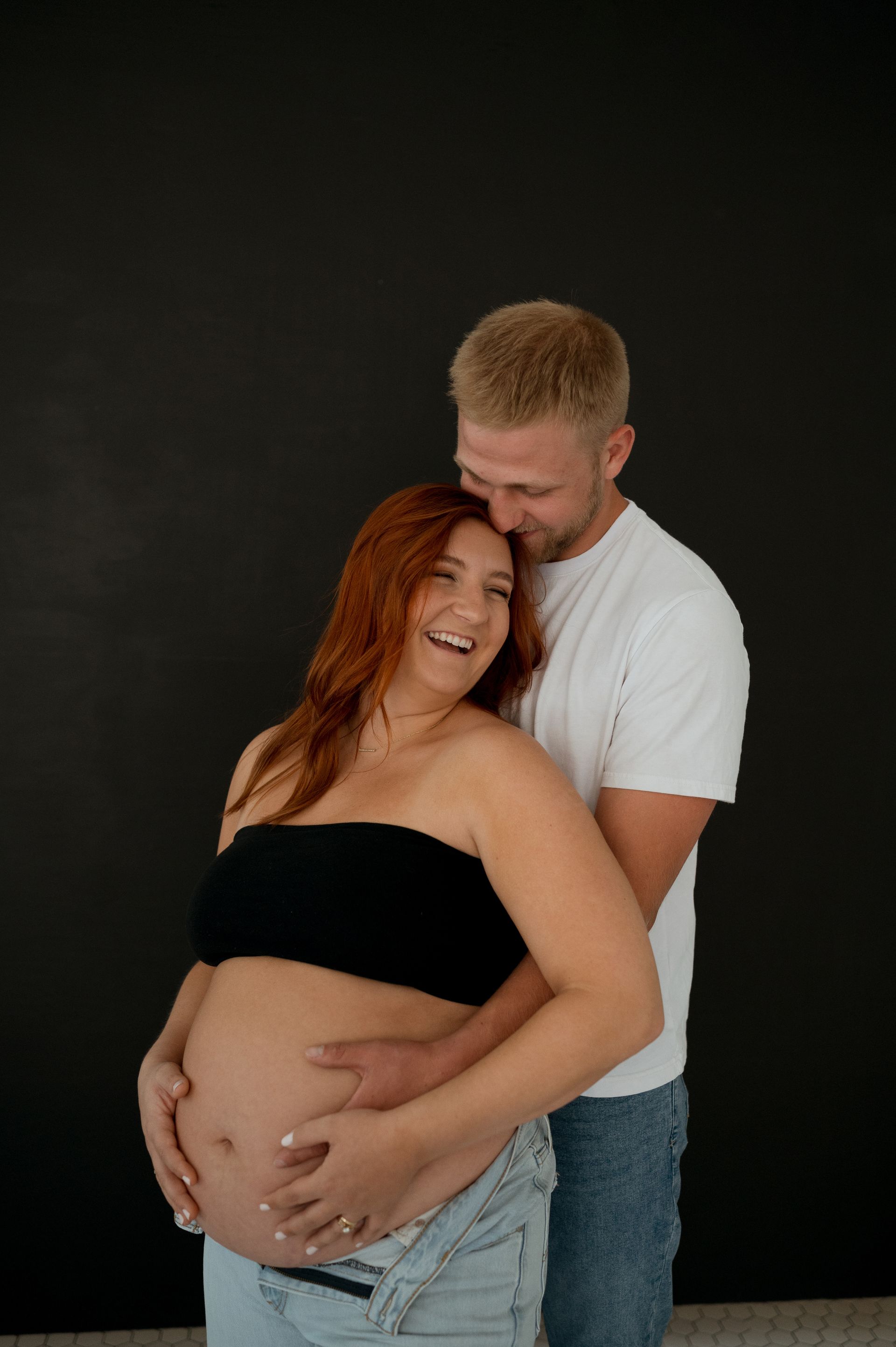 A man is hugging a pregnant woman in front of a black wall.