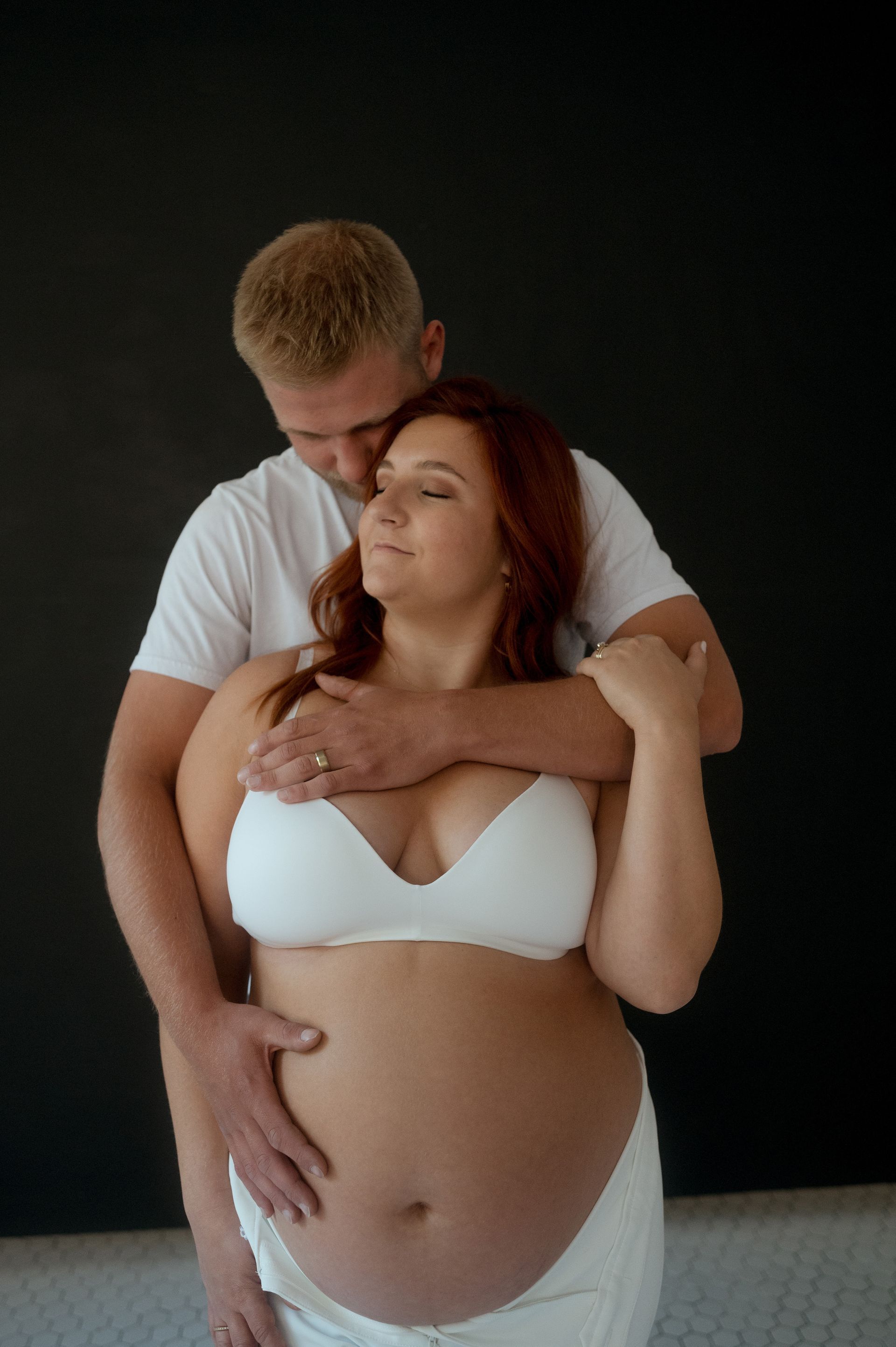 A man is hugging a pregnant woman in a white bra.