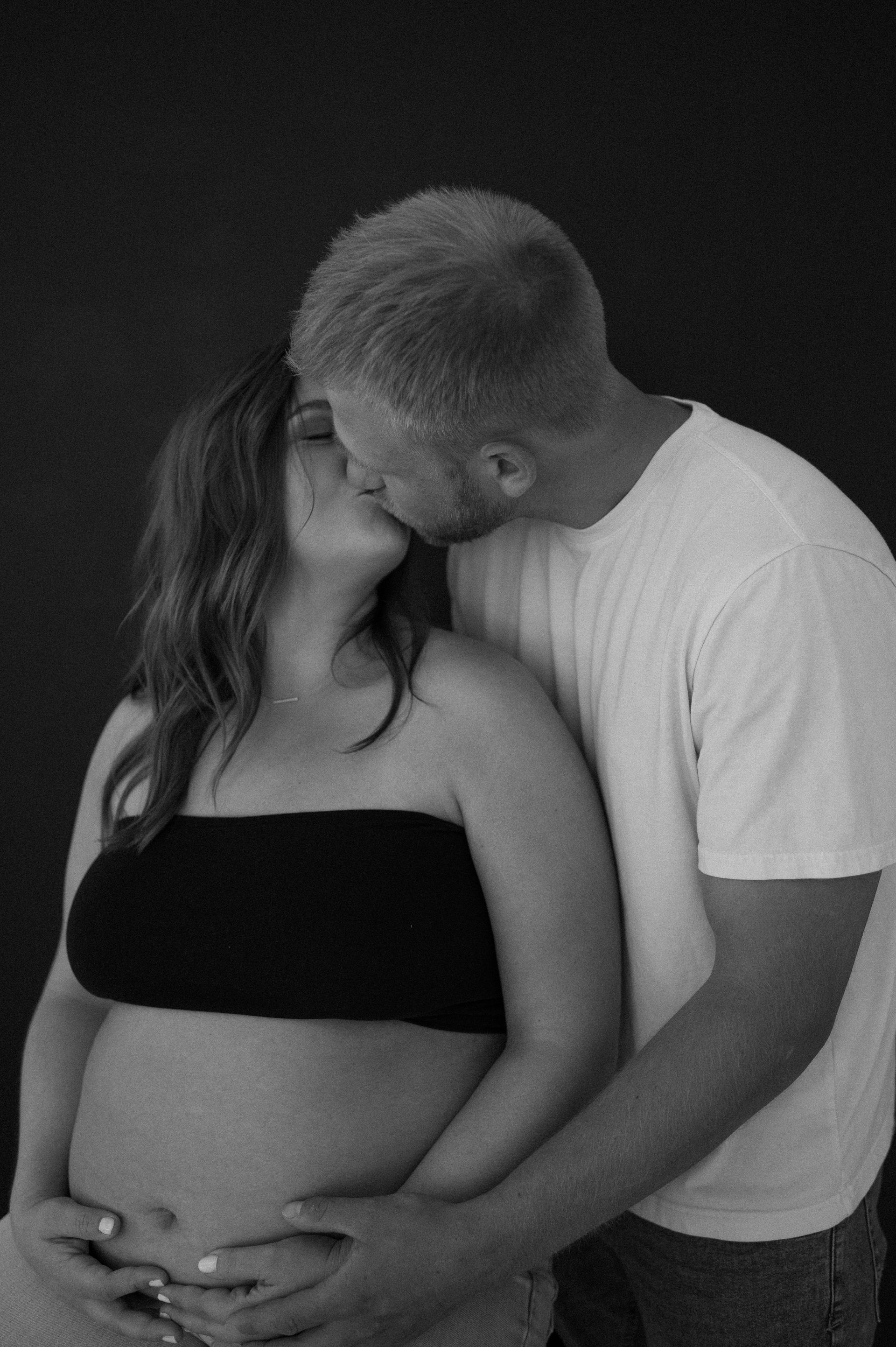 A man is kissing a pregnant woman on the cheek in a black and white photo.