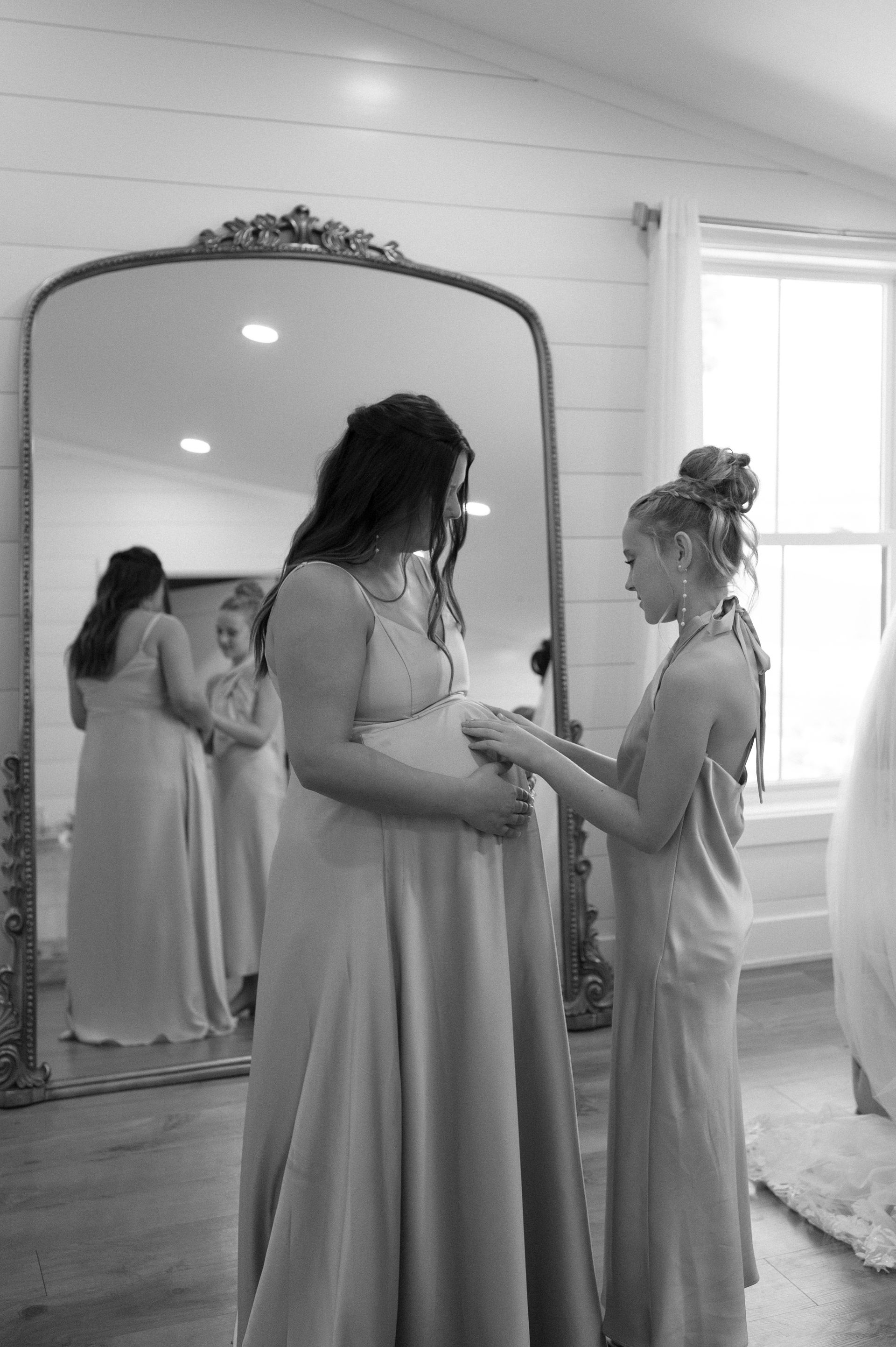 A pregnant woman and her bridesmaid are standing in front of a mirror.