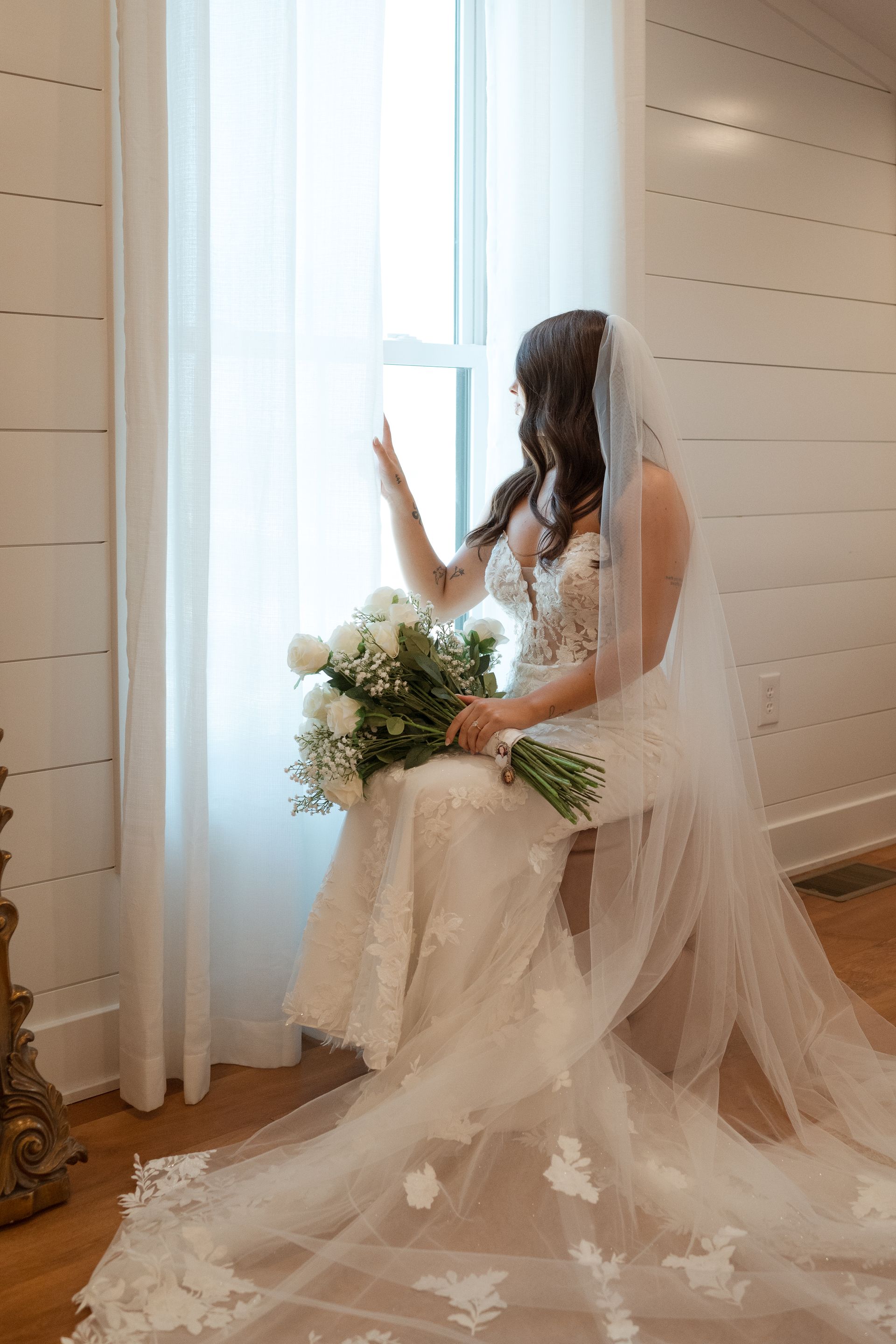 A bride in a wedding dress and veil is sitting in front of a window holding a bouquet of flowers.