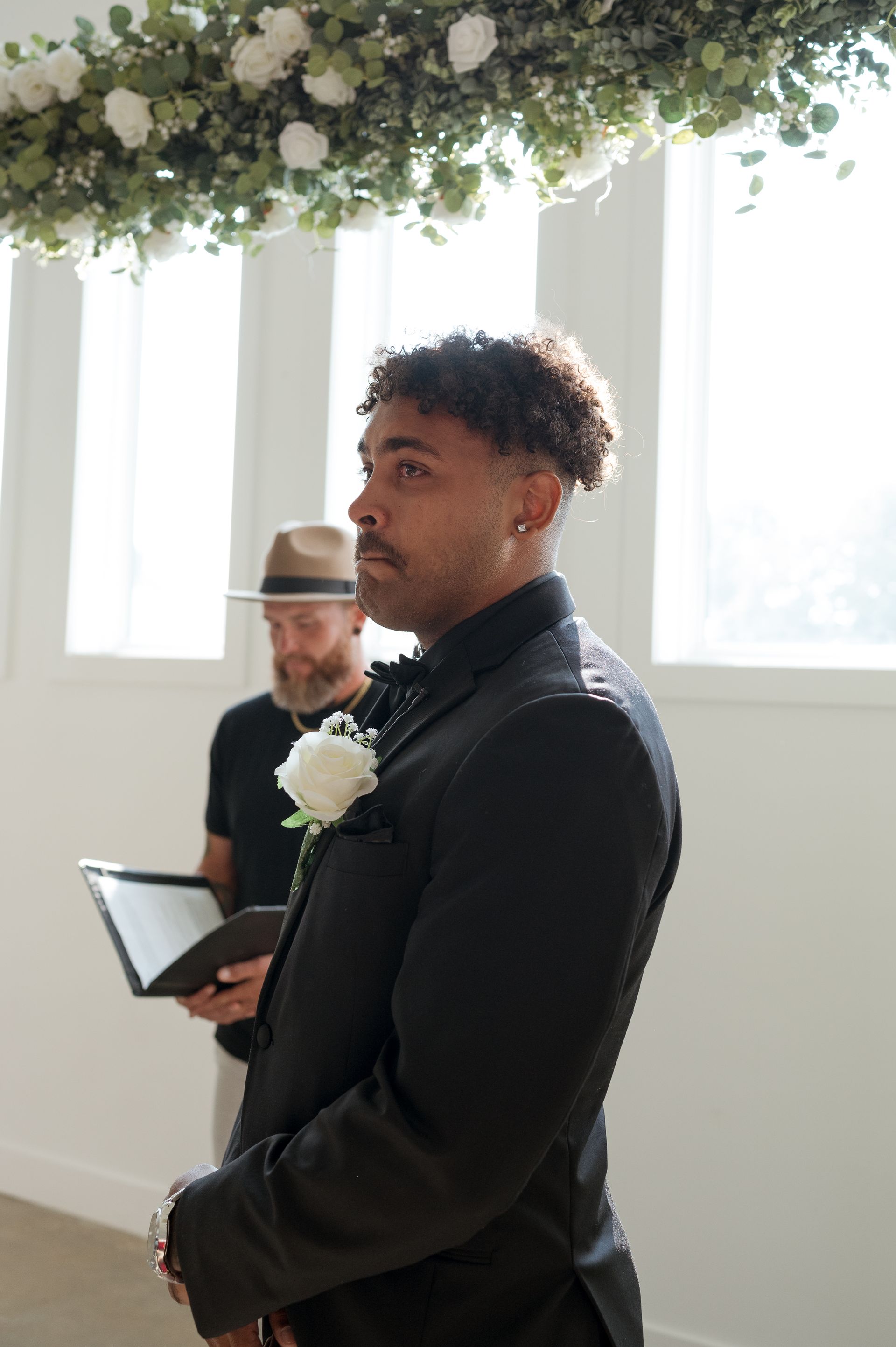 A man in a black suit is standing in front of a window holding a book.