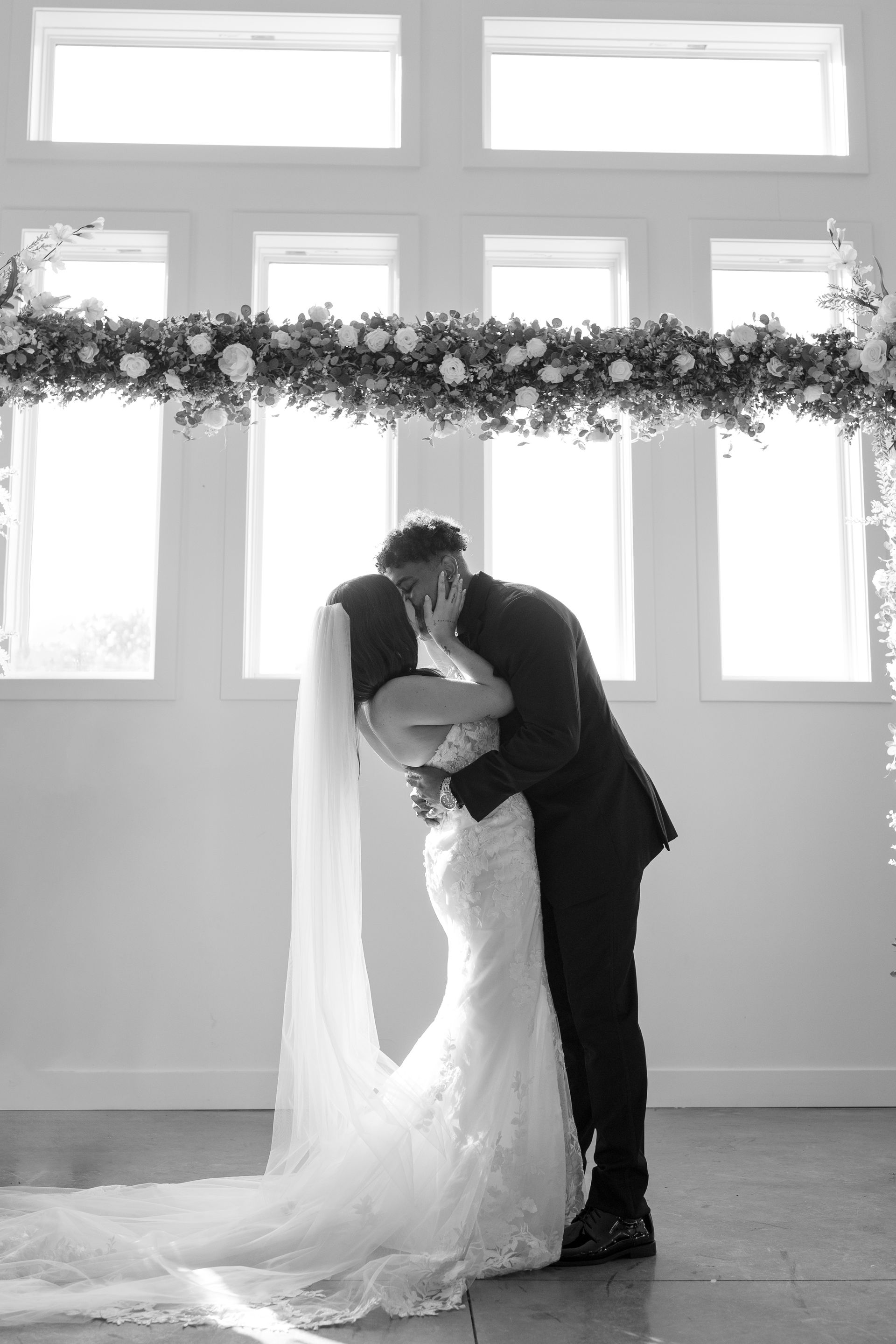 A bride and groom are kissing under a floral arch at their wedding.