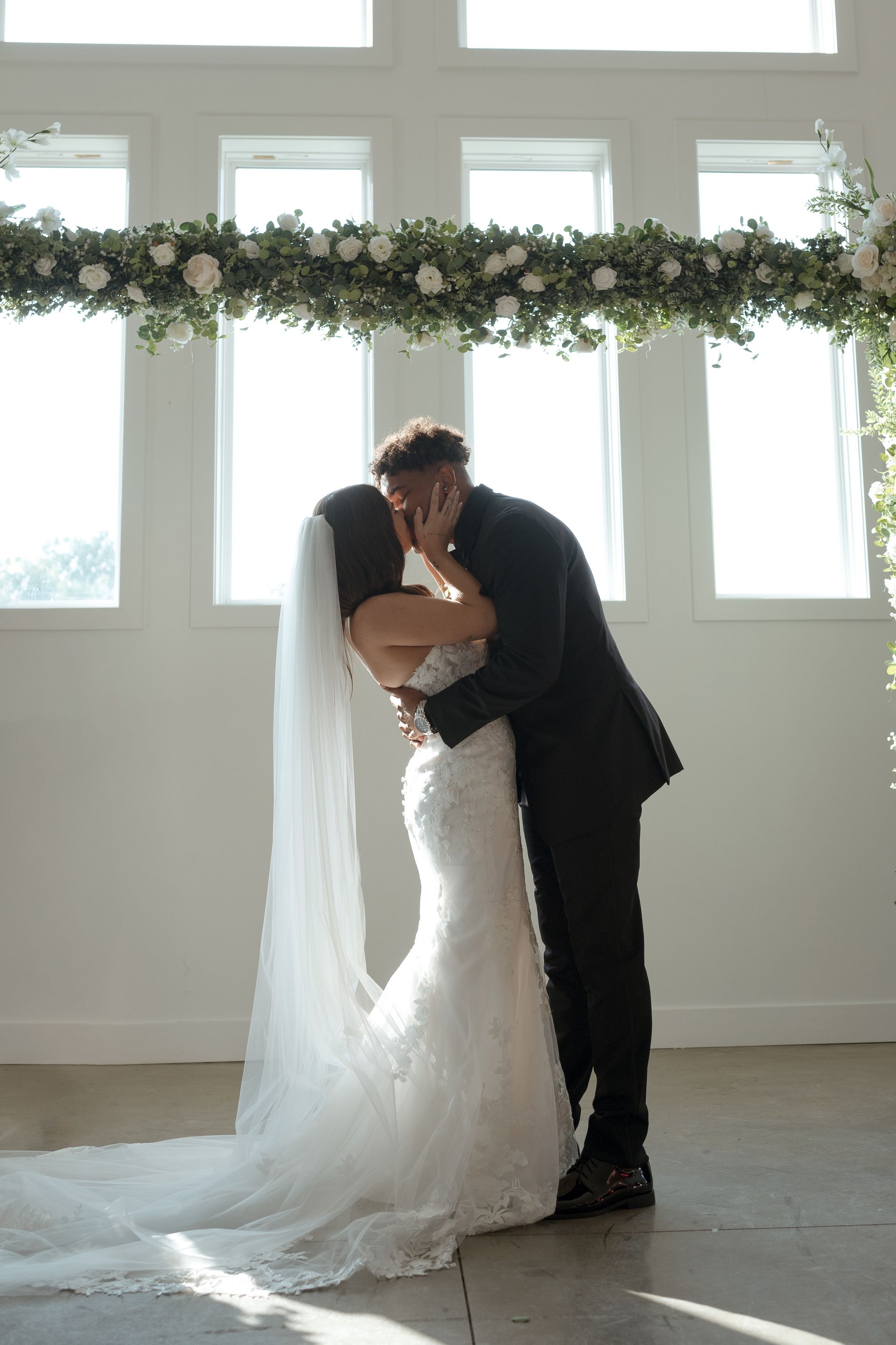 A bride and groom are kissing under a floral arch at their wedding ceremony.