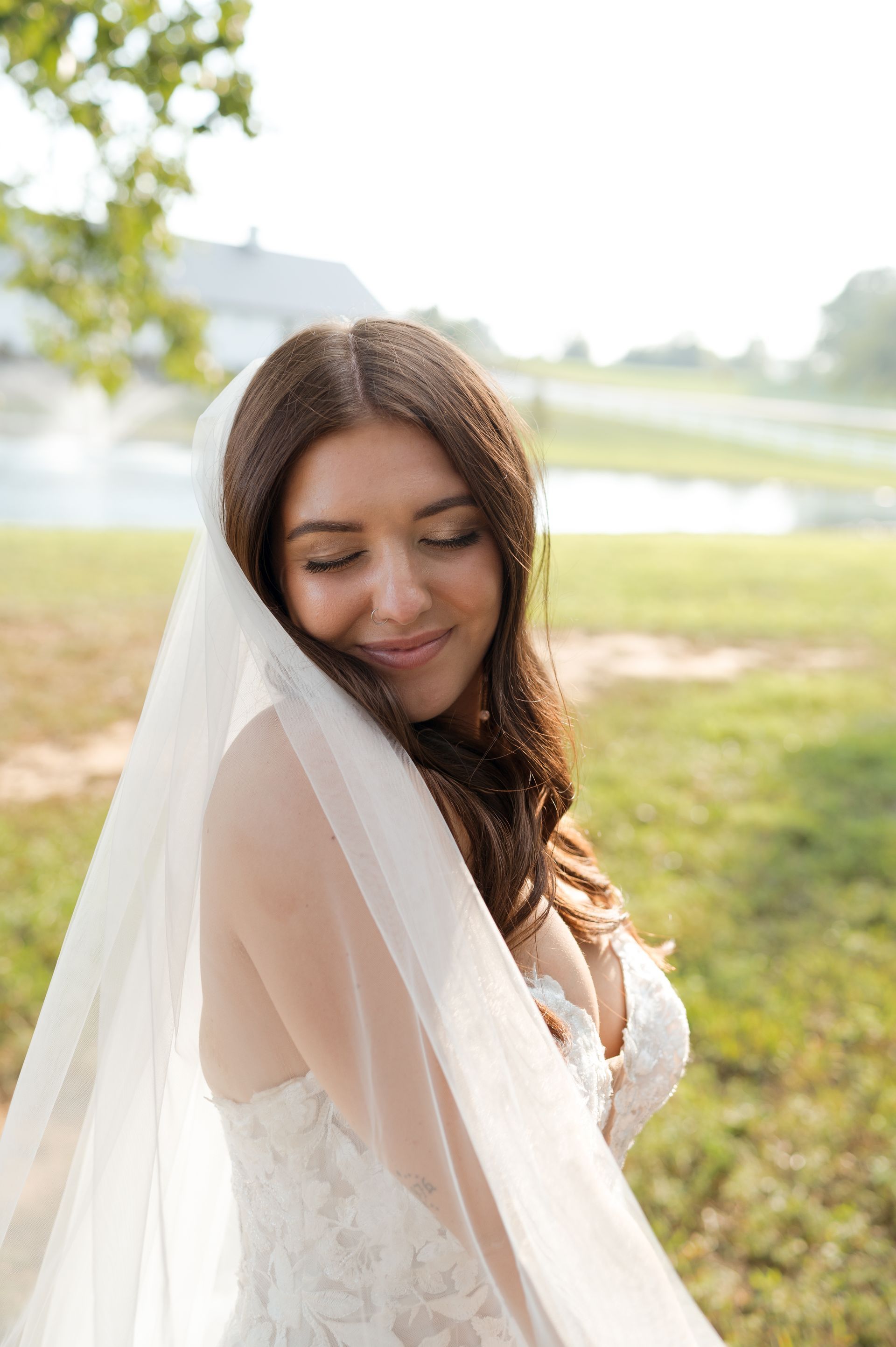 A bride in a wedding dress and veil is standing in a field with her eyes closed.