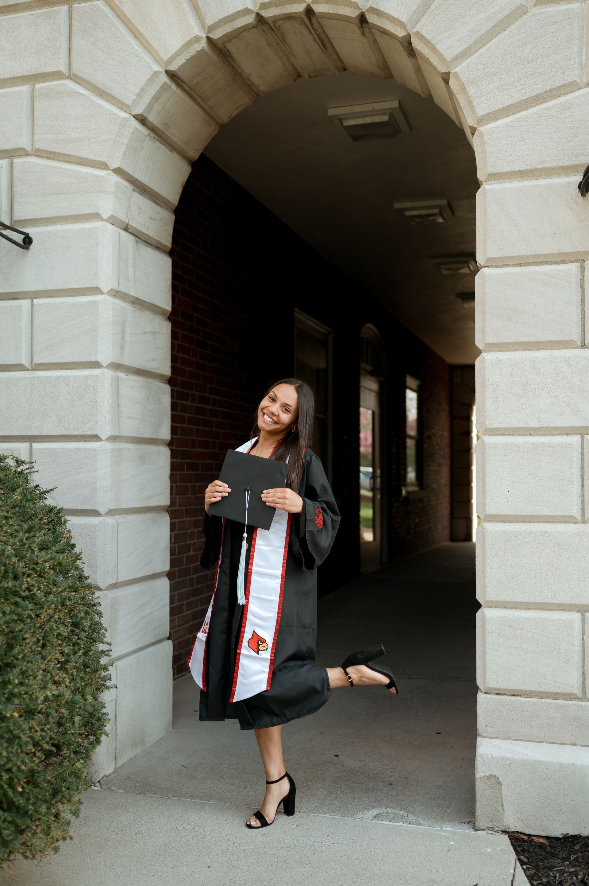 A woman in a graduation cap and gown is standing in front of a building holding a diploma.