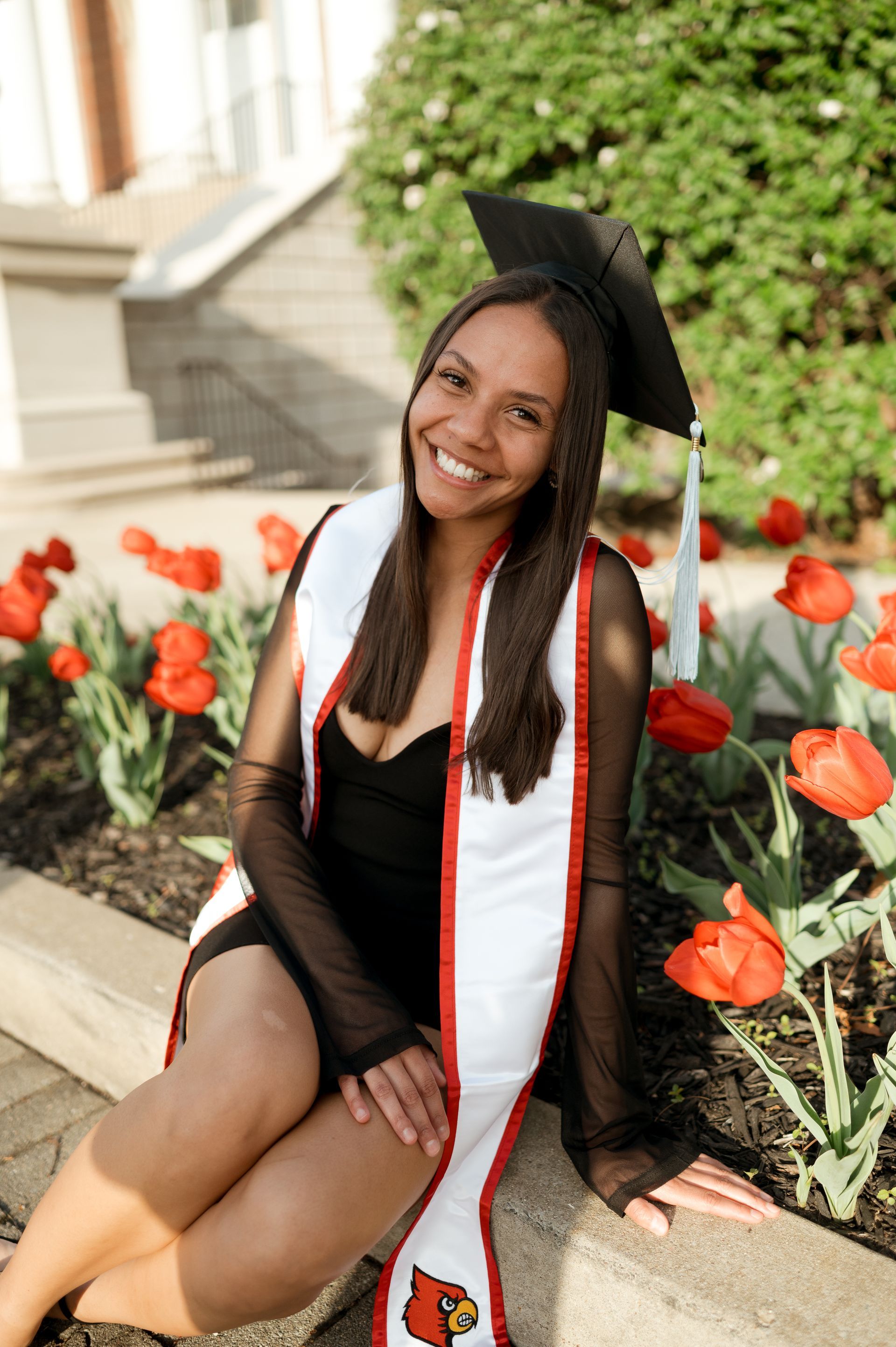 A woman in a graduation cap and gown is sitting in front of flowers.