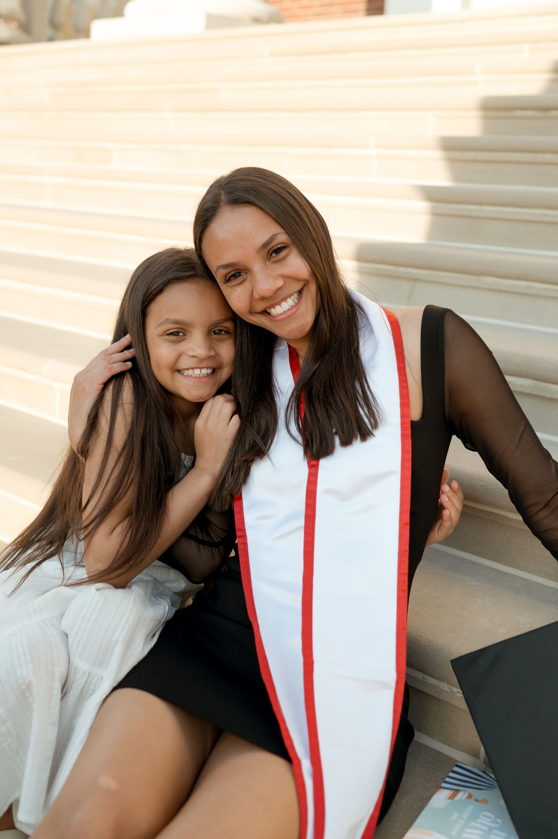 A woman in a graduation cap and gown is sitting next to a little girl.