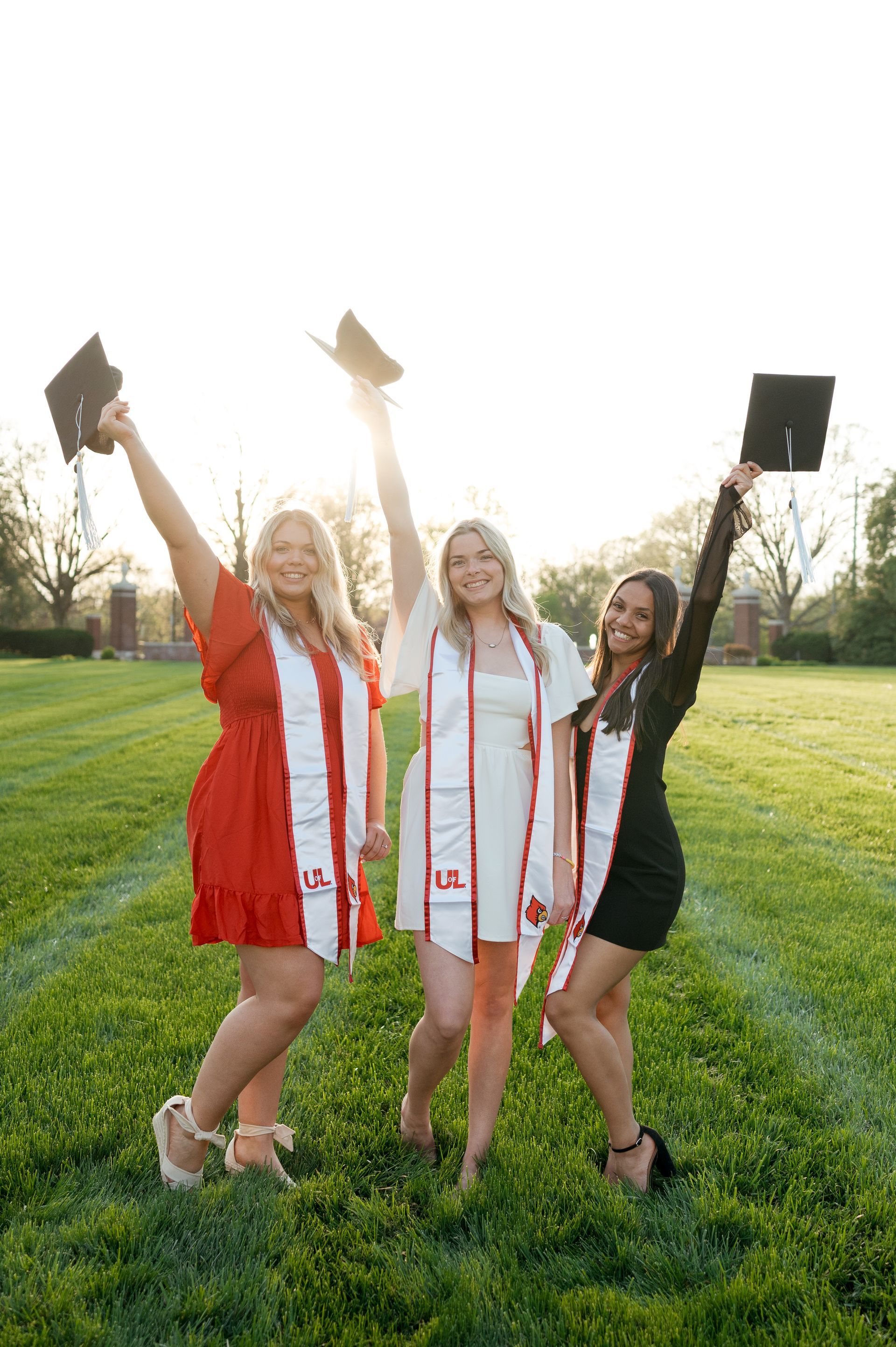 Three female graduates are standing in a field holding their caps in the air.