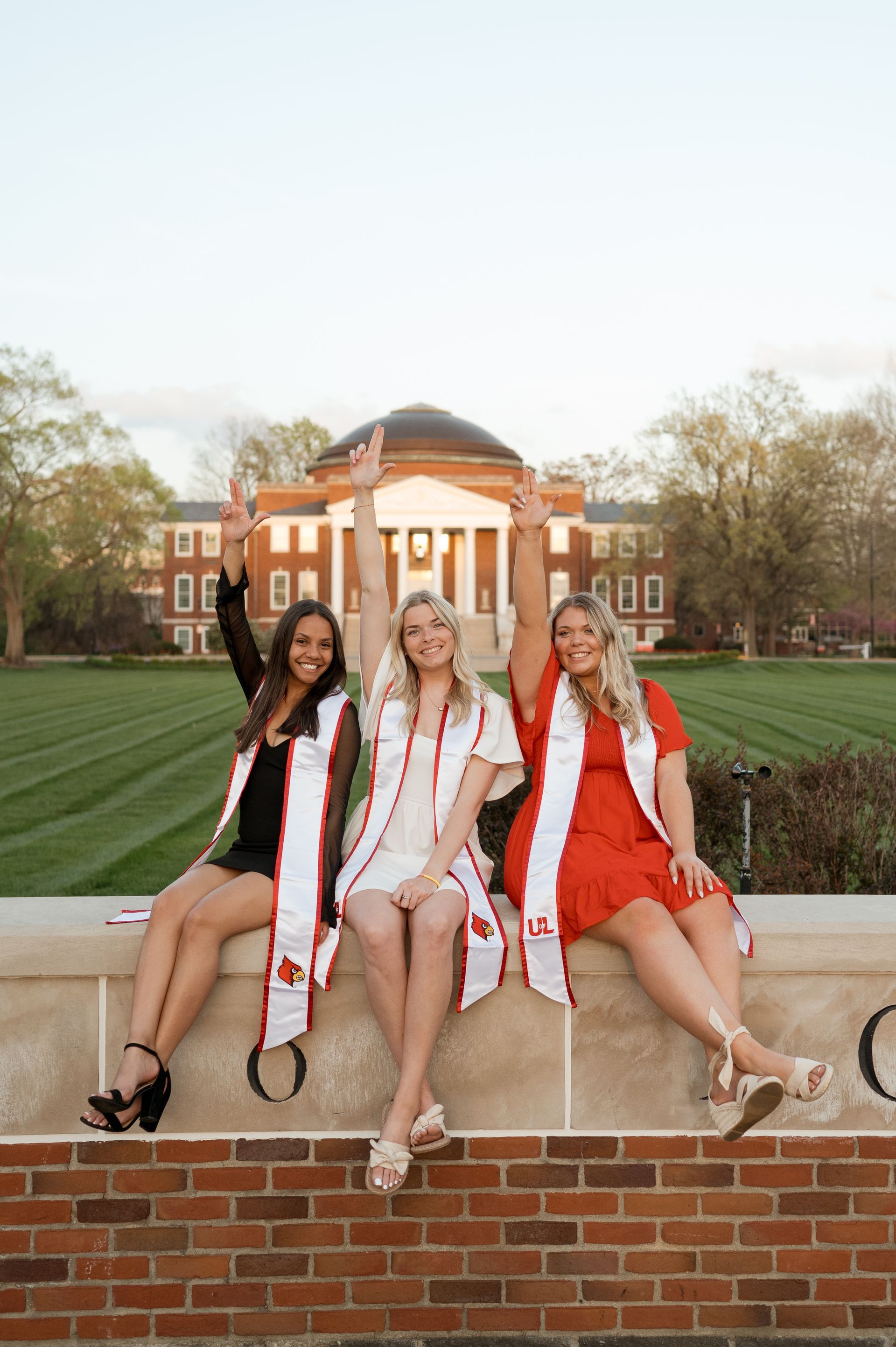 Three women are sitting on a brick wall with their arms in the air.