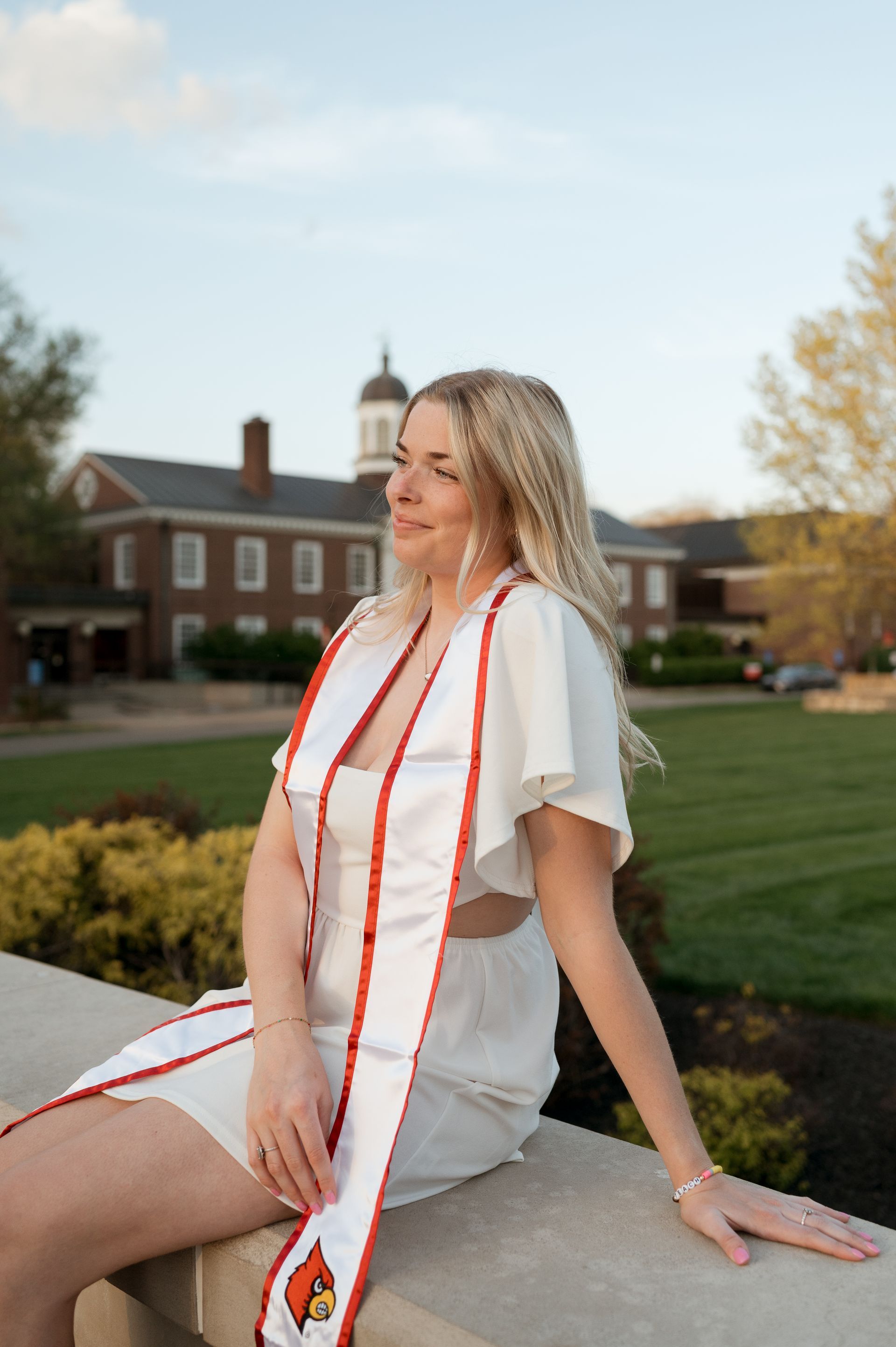 A woman in a white dress and red sash is sitting on a wall.