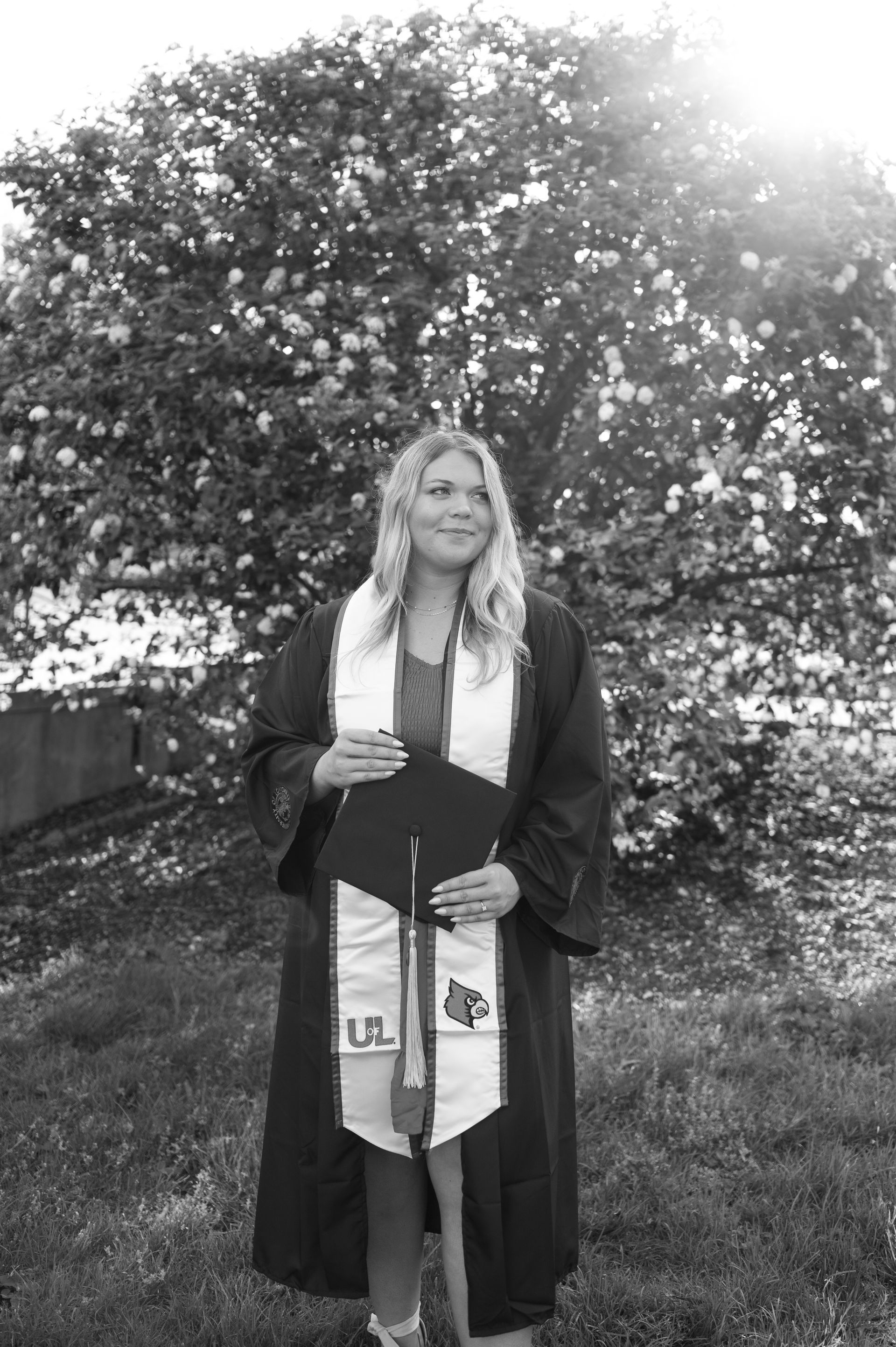 A woman in a graduation cap and gown is standing in front of a tree.