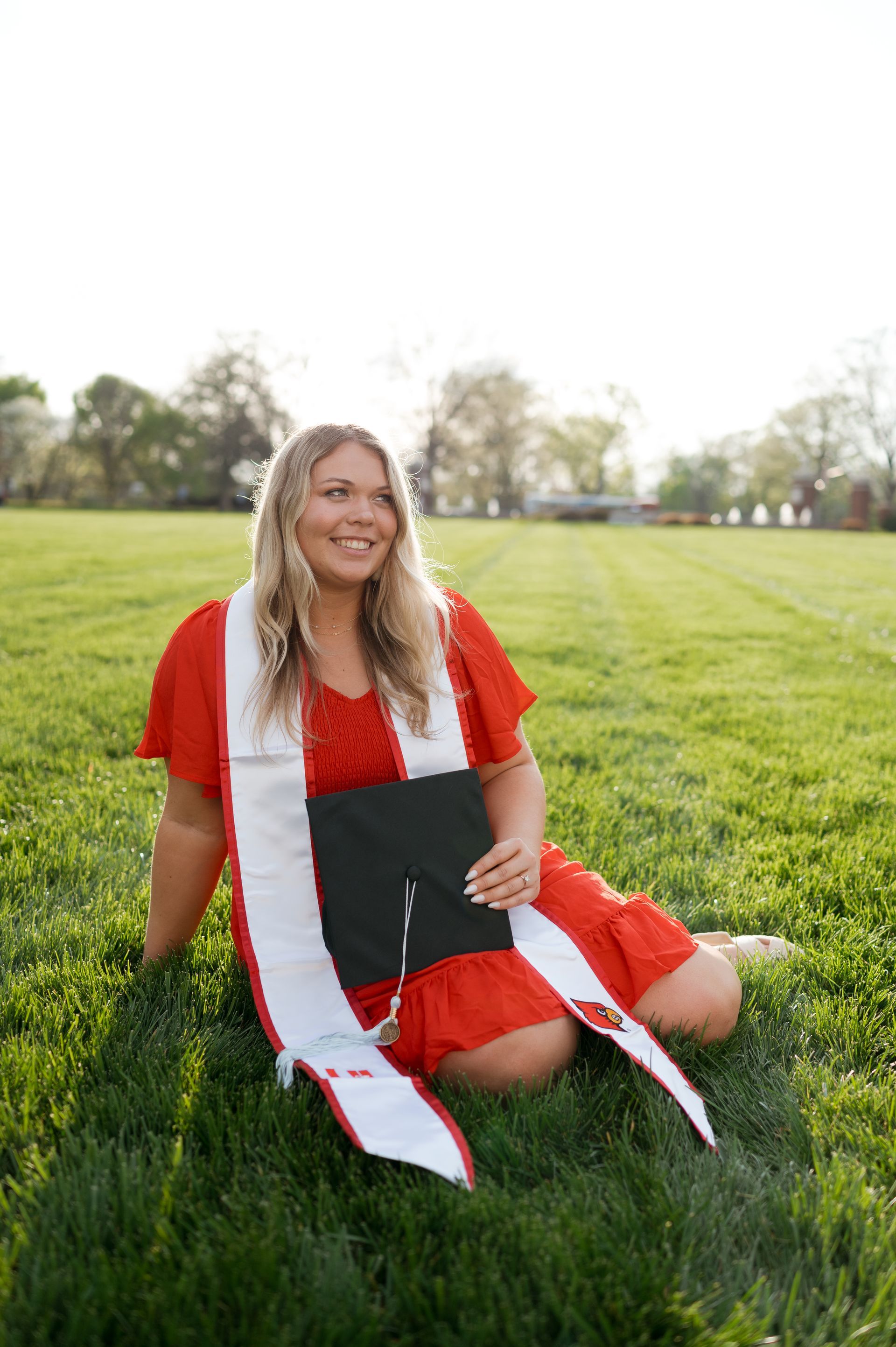A woman in a red dress is sitting in the grass holding a graduation cap.