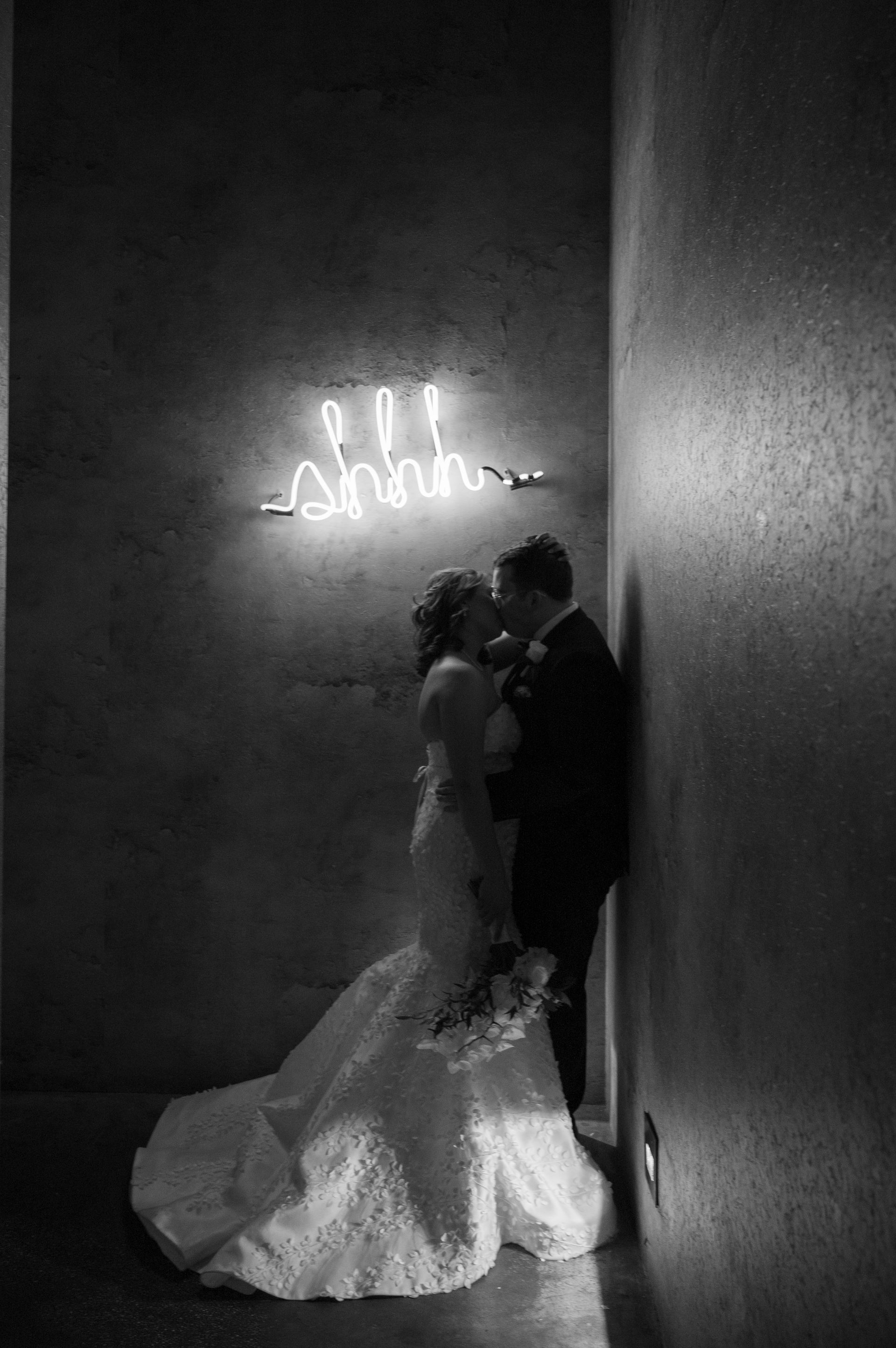 A bride and groom are kissing in front of a neon sign.