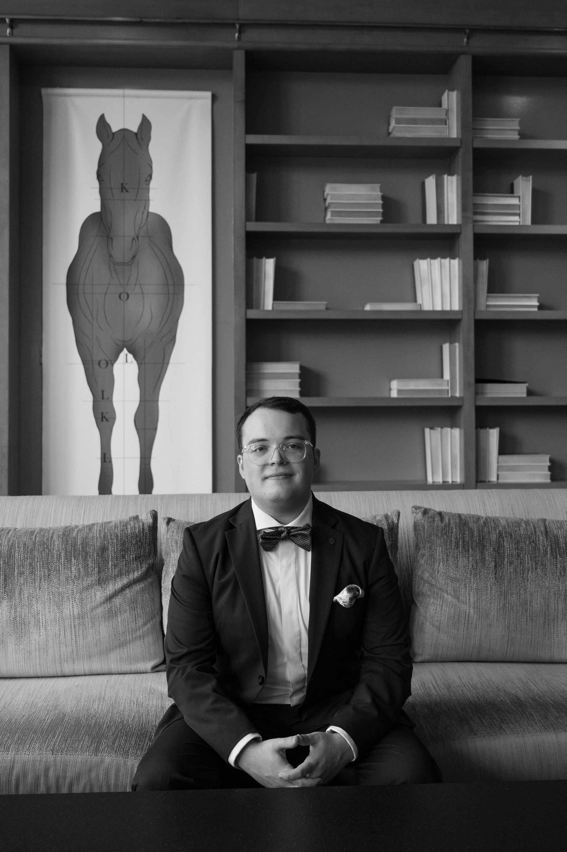 A man in a tuxedo is sitting on a couch in front of a picture of a horse.