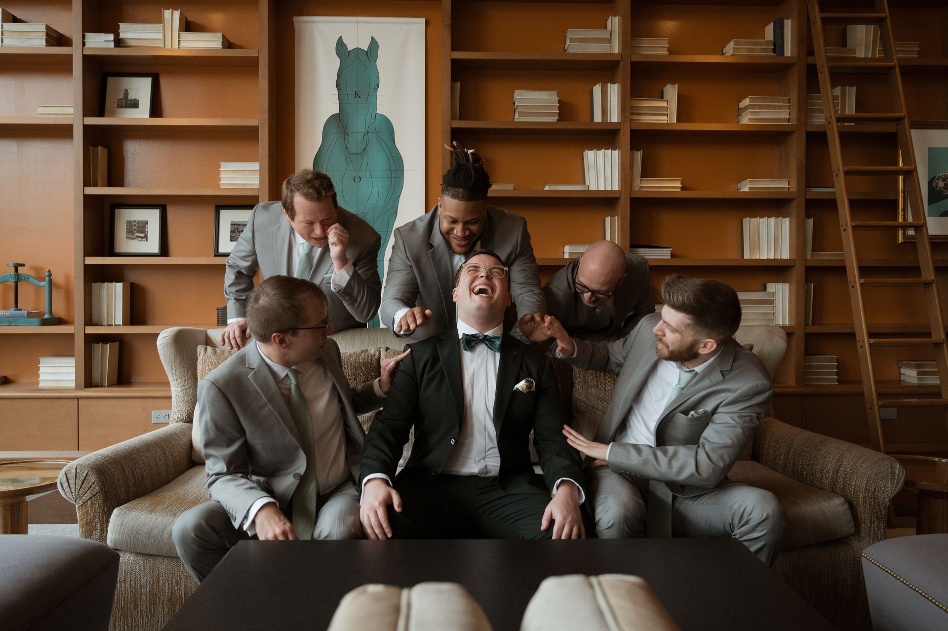 A group of men in suits are sitting around a man in a tuxedo on a couch.