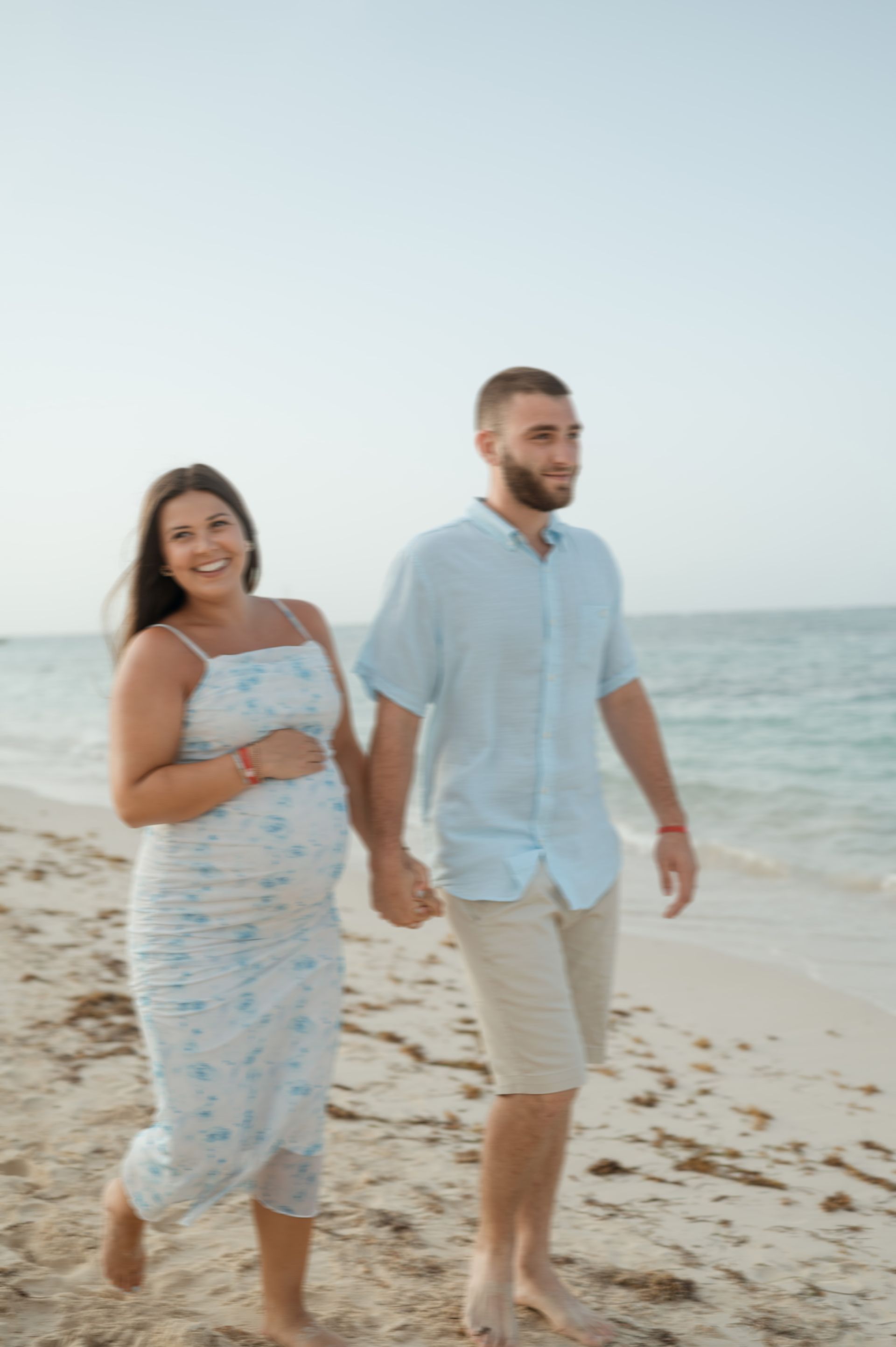 A man and a pregnant woman are walking on the beach holding hands.