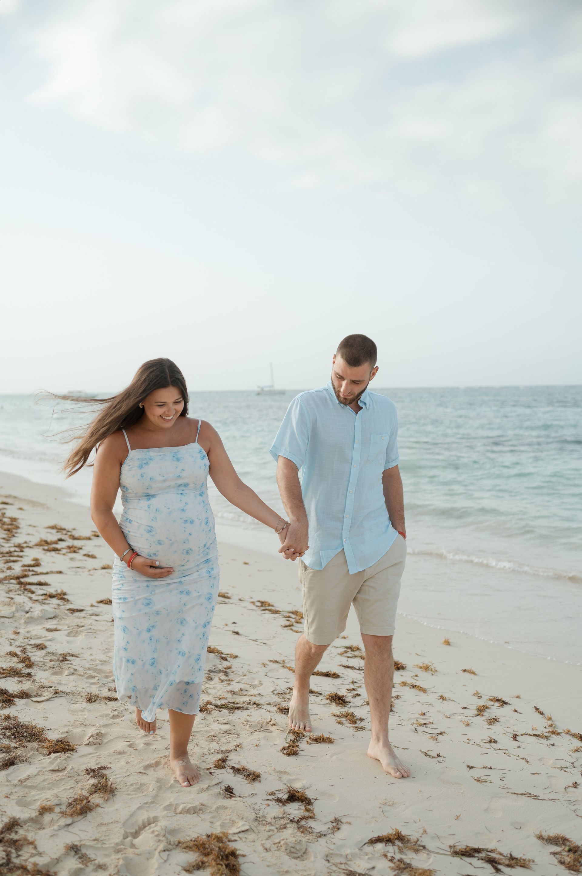 A man and a pregnant woman are walking on the beach holding hands.