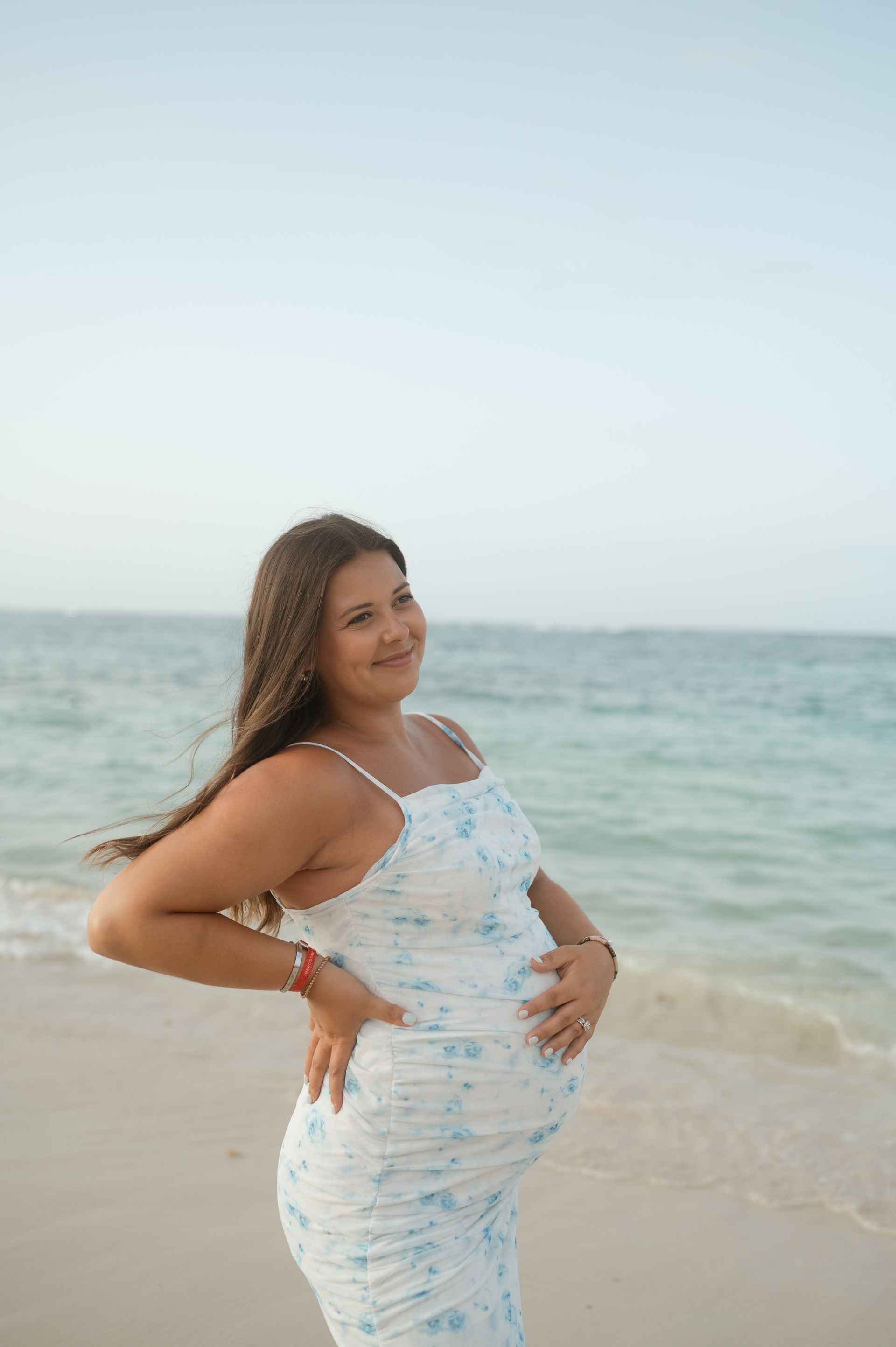 A pregnant woman is standing on the beach holding her belly.