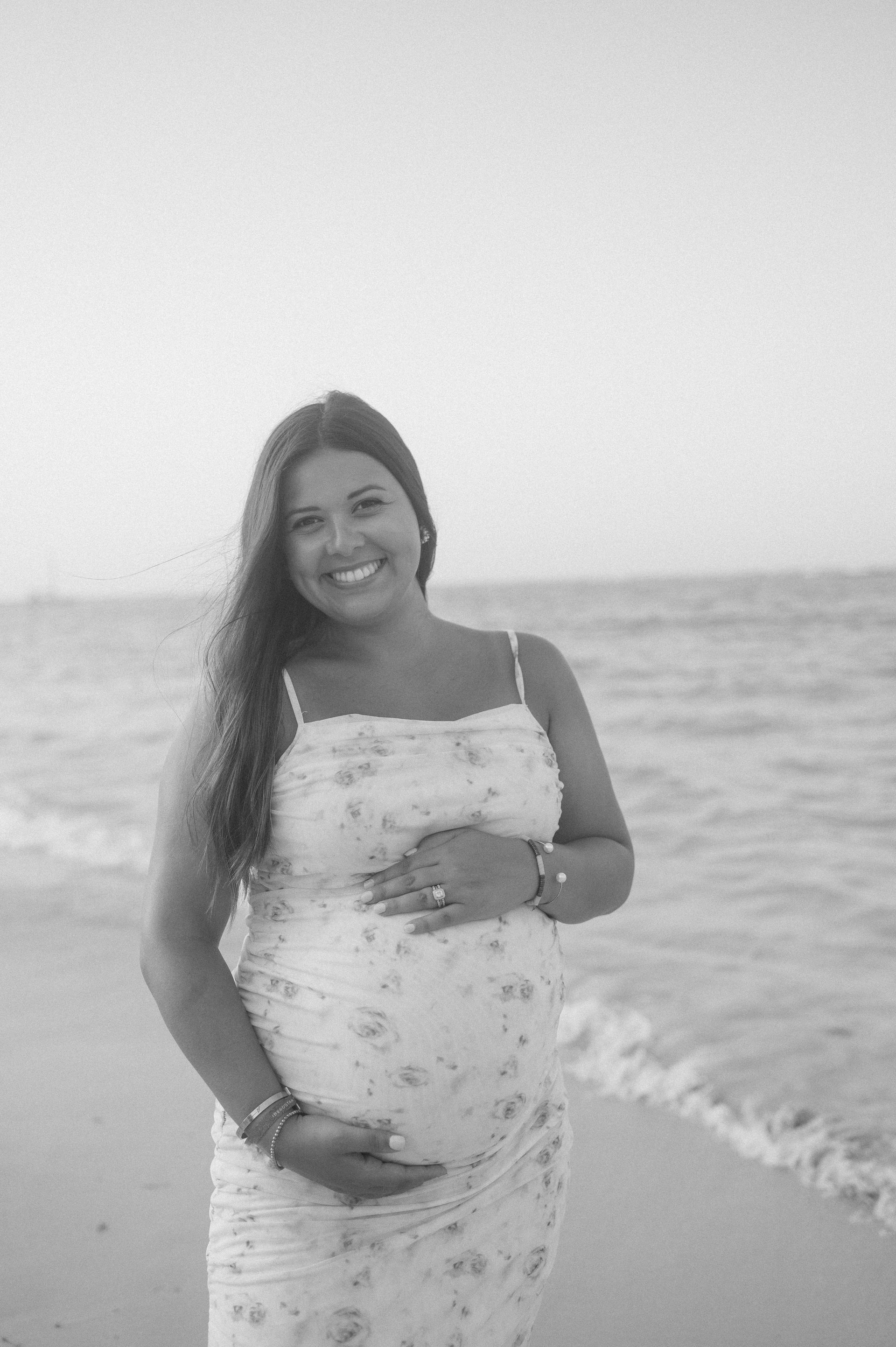 A pregnant woman is standing on the beach holding her belly and smiling.
