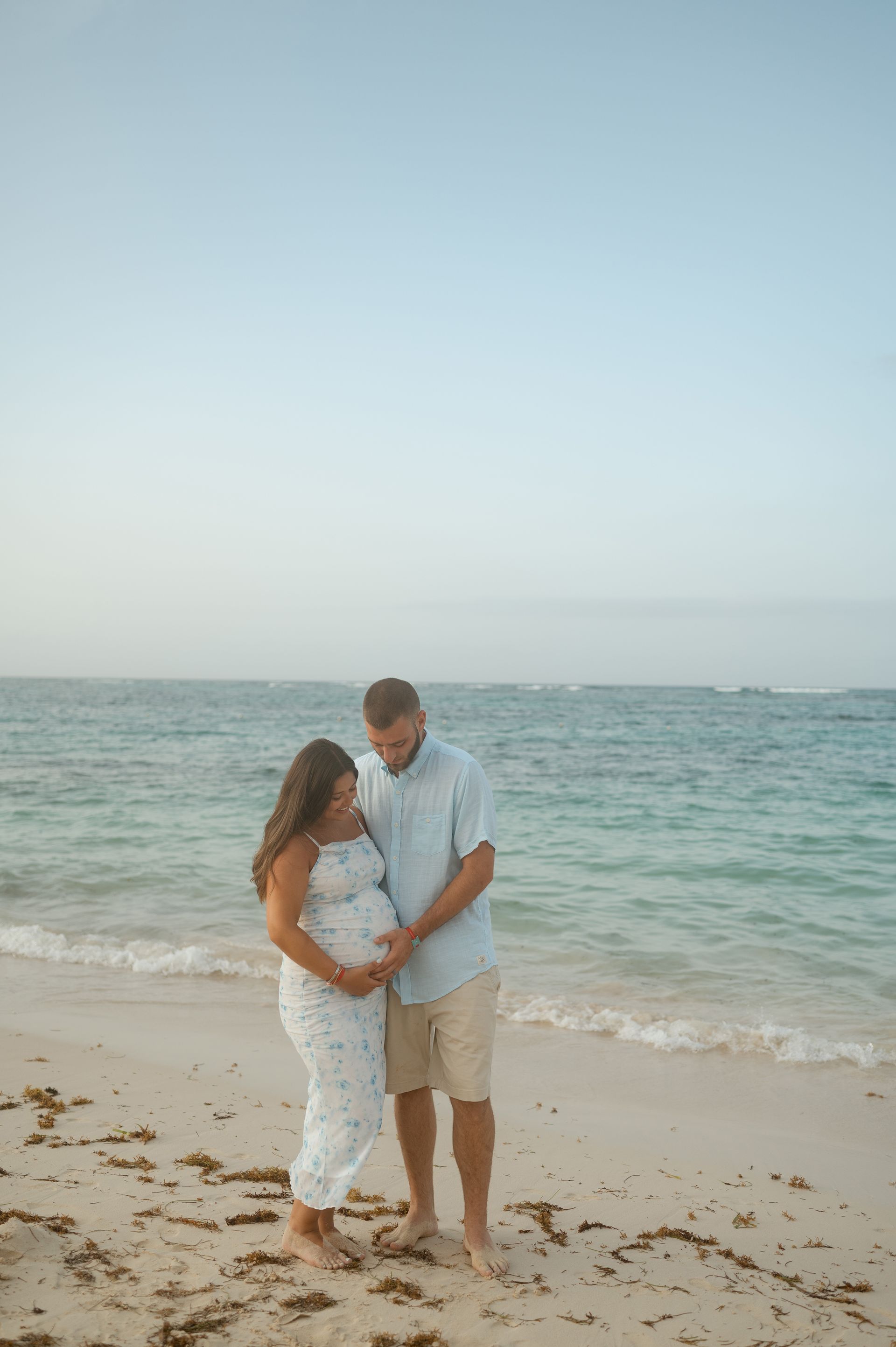 A man and a pregnant woman are standing on a beach holding hands.