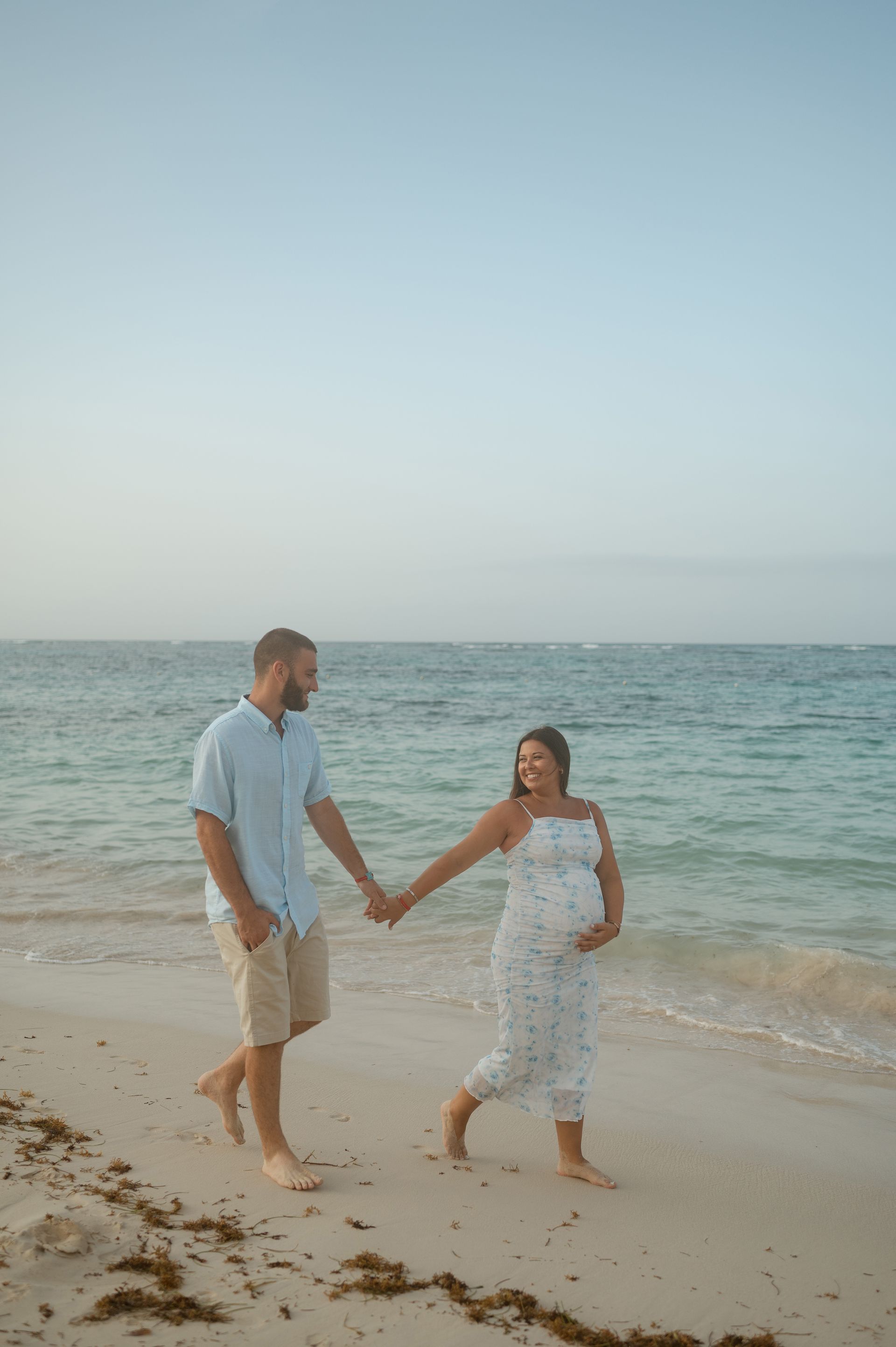 A man and a pregnant woman are walking on the beach holding hands.