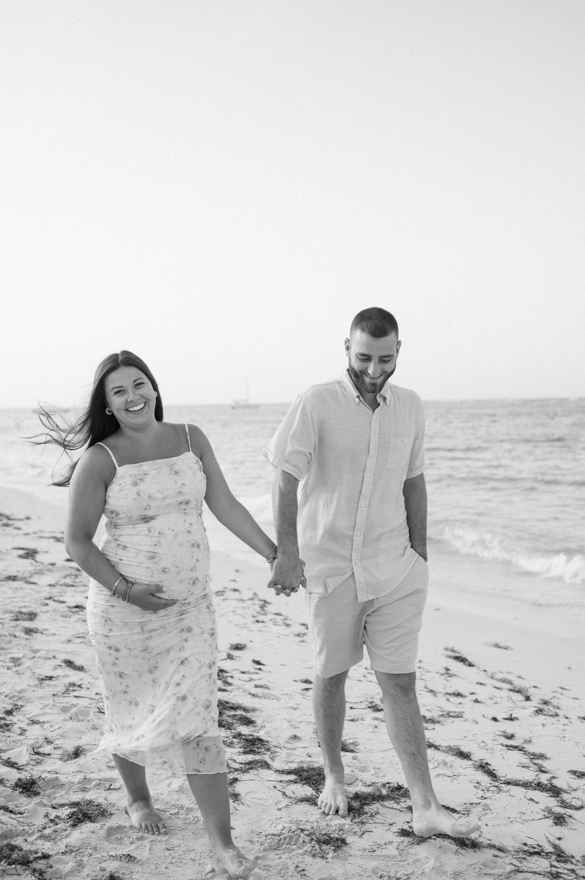 A man and a pregnant woman are walking on the beach holding hands.