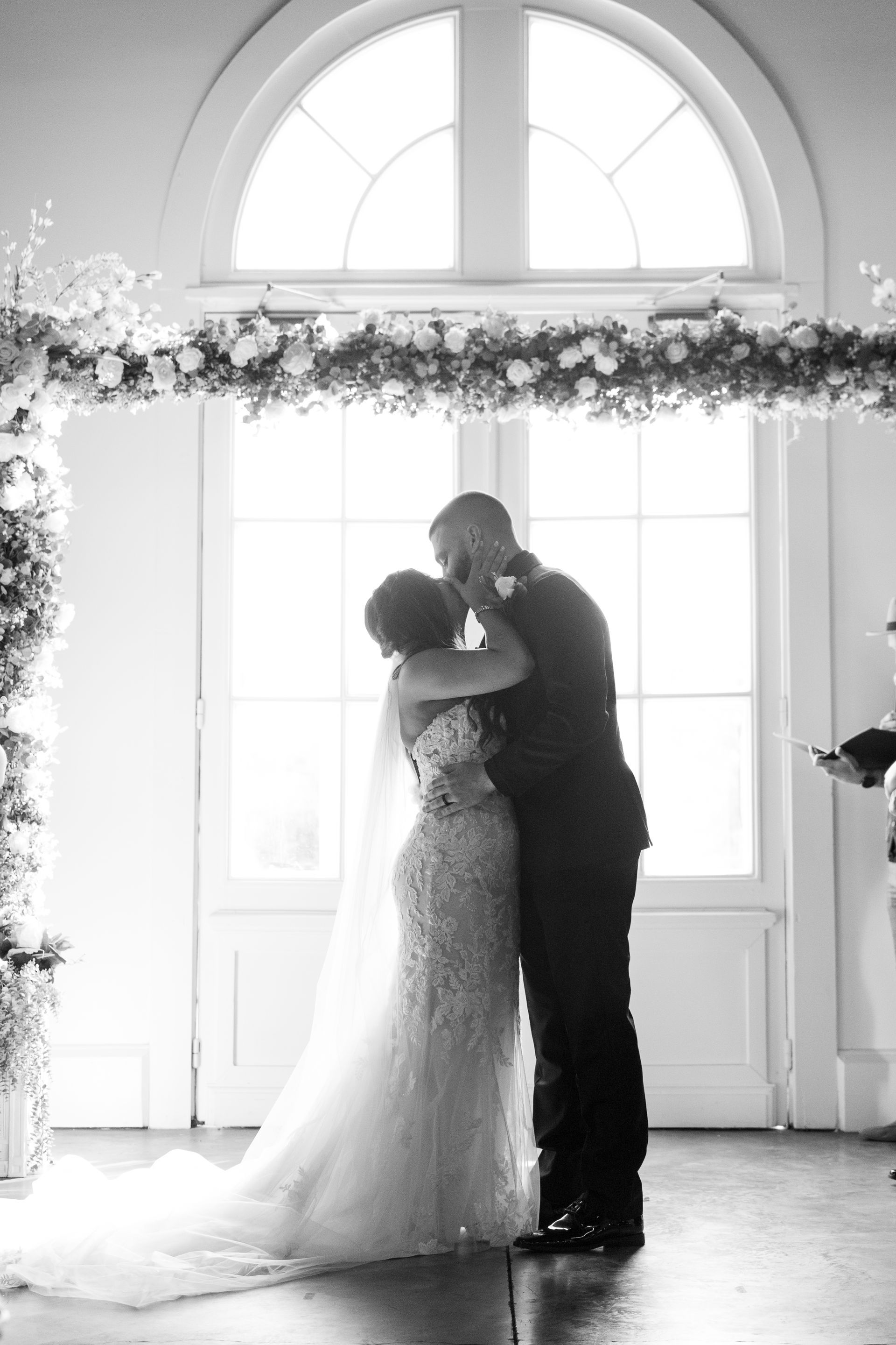 A black and white photo of a bride and groom kissing during their wedding ceremony.