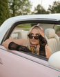 A woman wearing sunglasses and a scarf is sitting in a pink car.