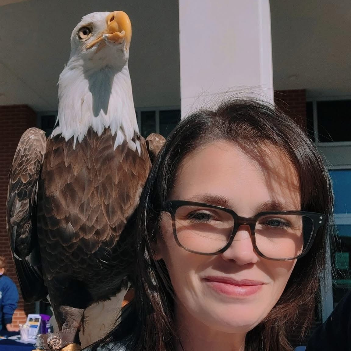 Woman with glasses smiles, bald eagle perched on her shoulder. Outdoor setting.
