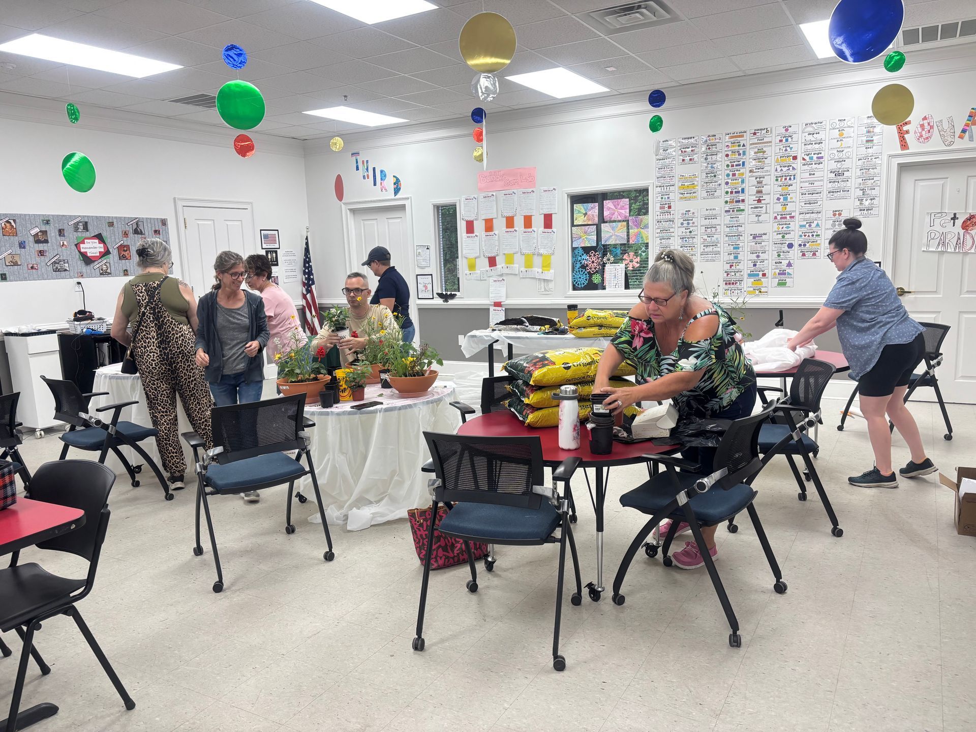 People seated at tables in a bright classroom or community room with colorful balloons and posters on the walls