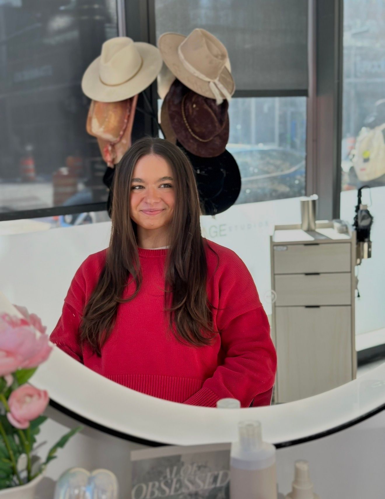 Woman with long brown hair in red sweater, reflected in a mirror. Hats hang behind her.