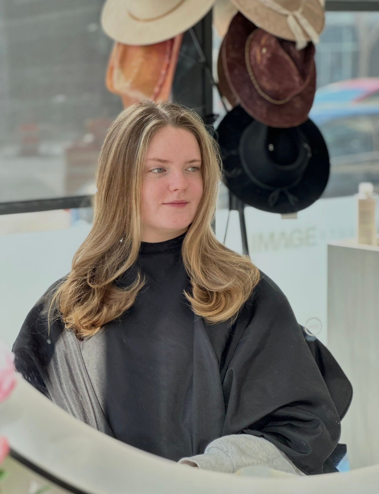 Woman with layered blonde hair at a salon wearing a black cape, with hats in the background.