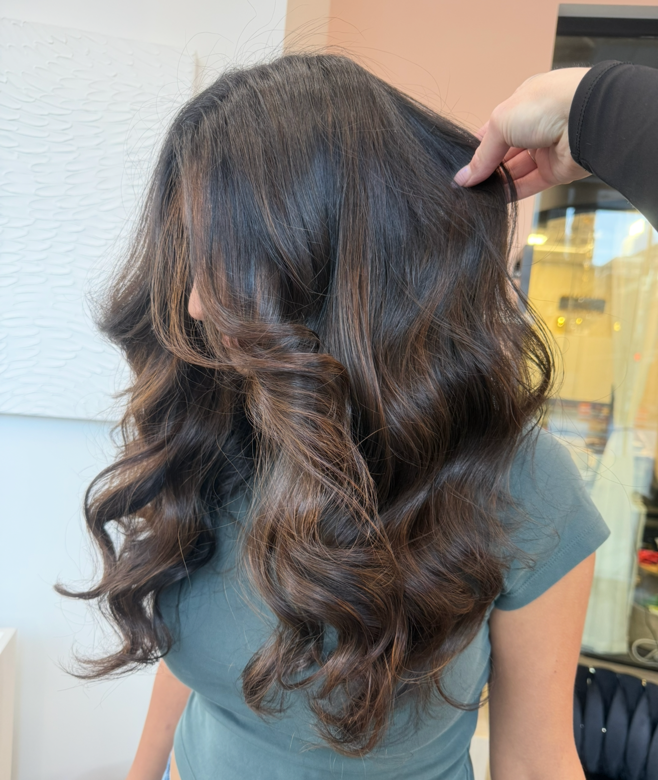 Woman with long, wavy brown hair being styled in a salon, hand touching the hair.