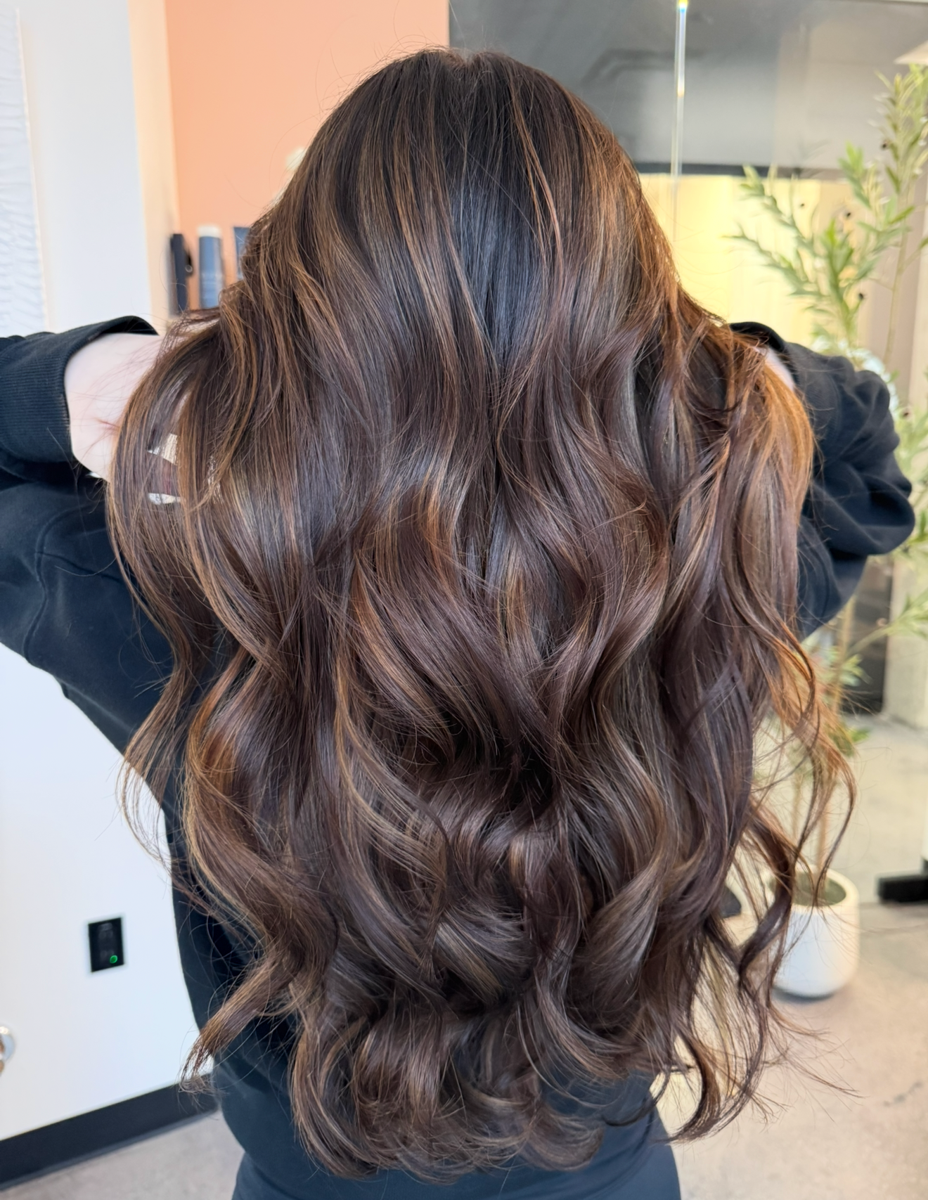 Woman with long, wavy brown hair with highlights, holding hair, indoors.