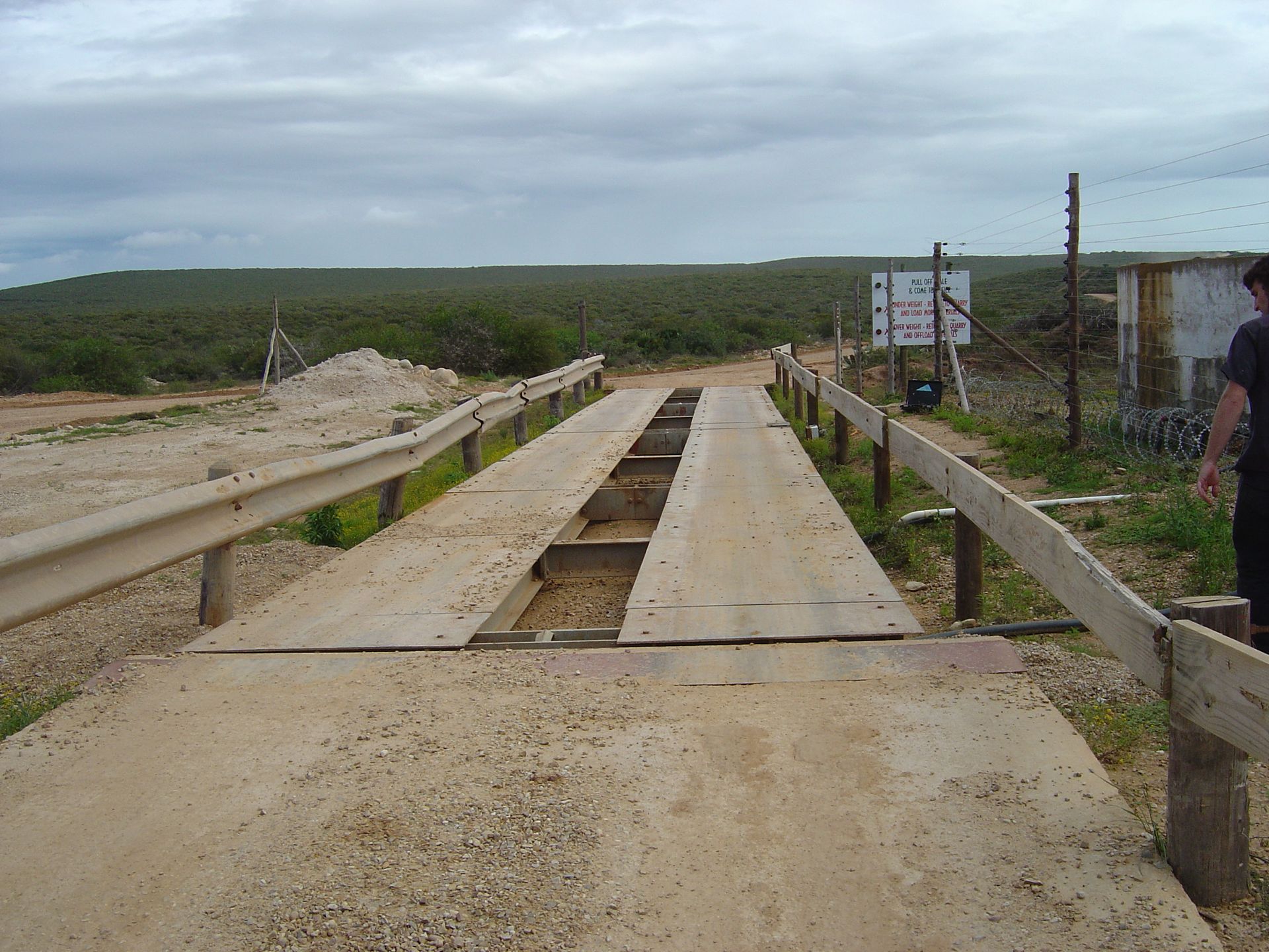 A man stands on a dirt road next to a wooden fence