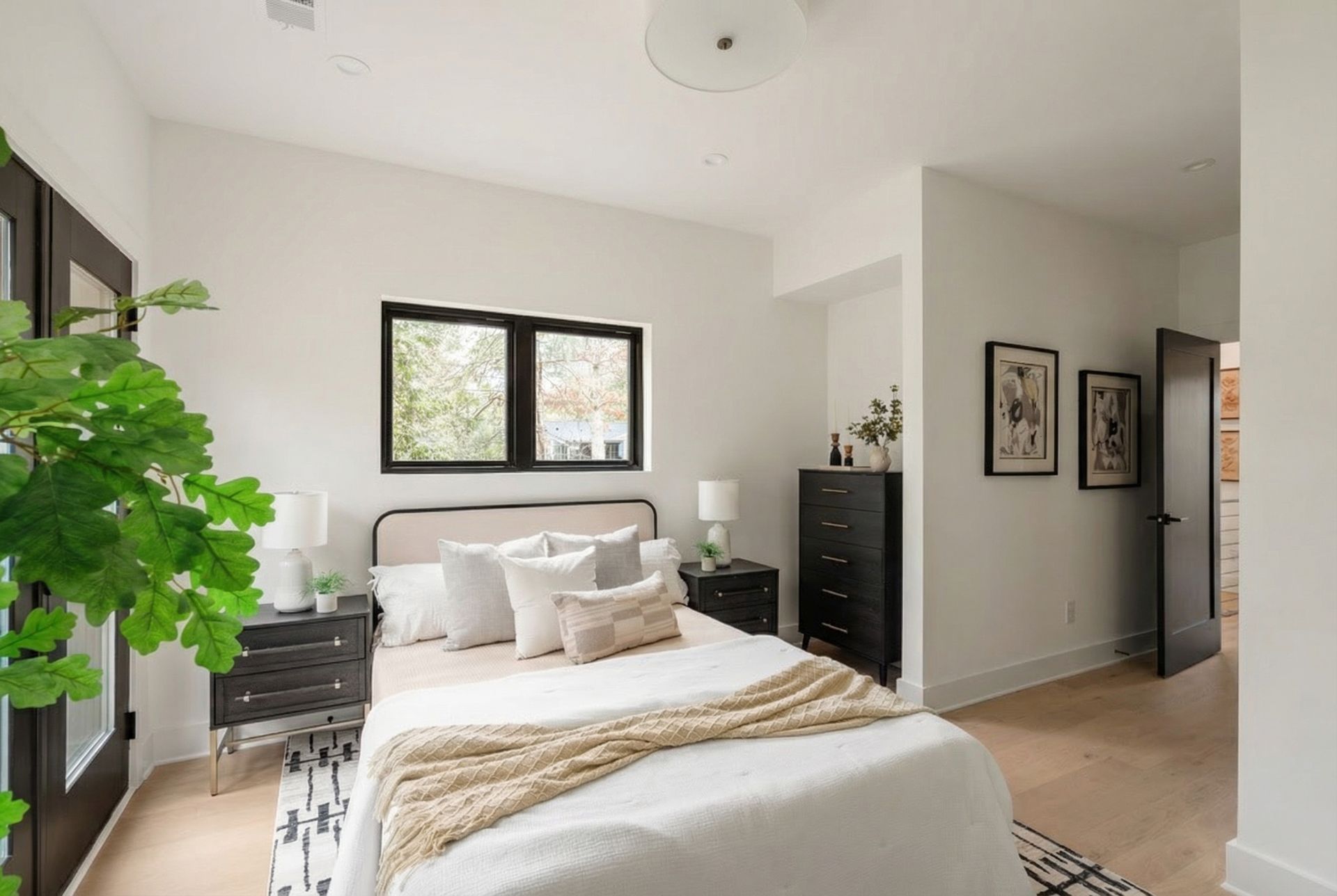 A modern, bright bedroom with a queen bed, black nightstands, a black dresser, and a large plant in the foreground.