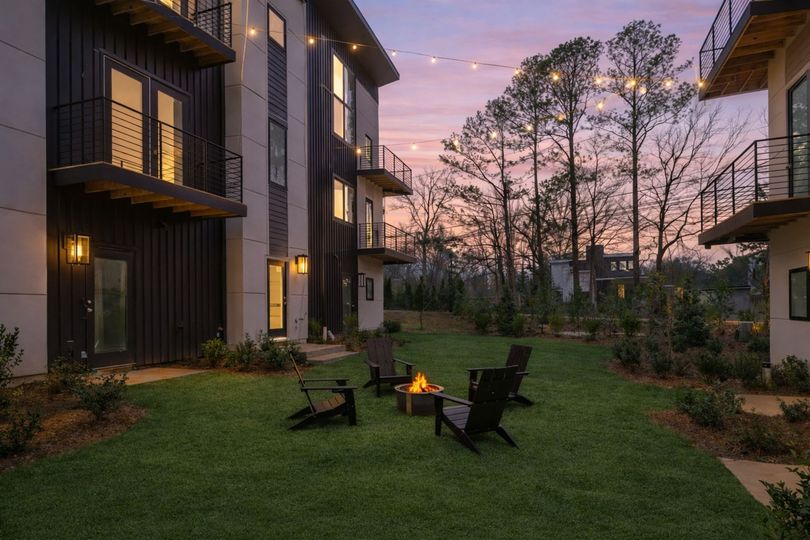 An outdoor courtyard at dusk with a fire pit, Adirondack chairs, and string lights between modern apartment buildings.