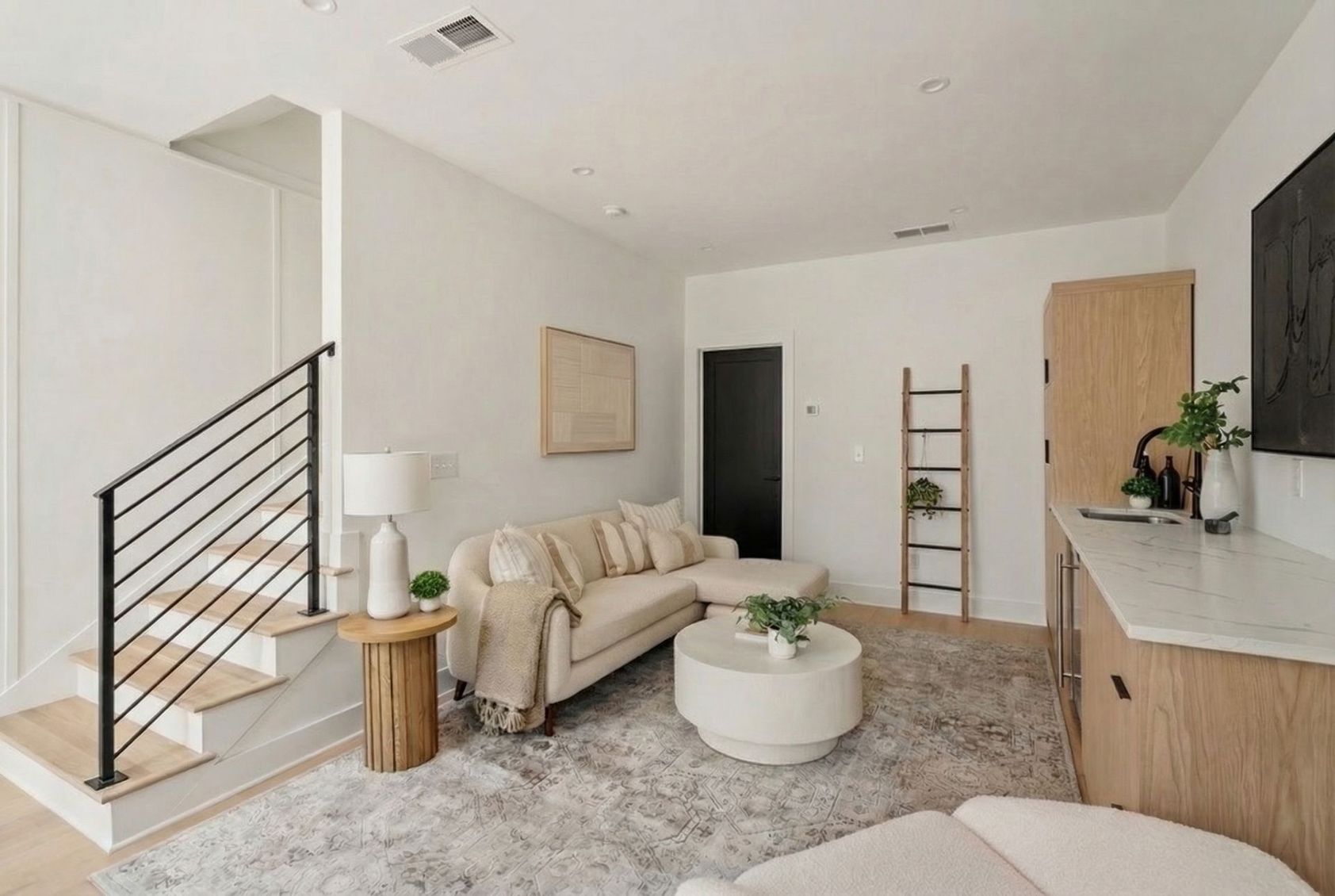 A modern living room with a neutral sectional sofa, round coffee table, staircase with black railing, and a wet bar.