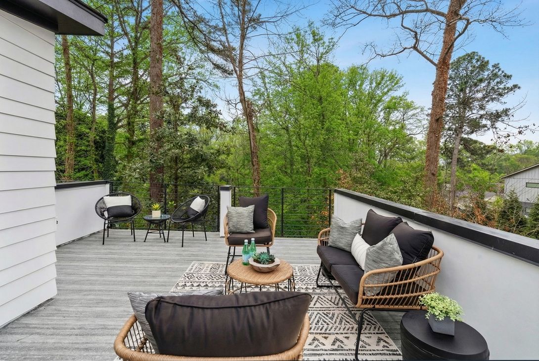 A modern, elevated wooden deck featuring a sofa, armchairs, and a small table, overlooking a lush green forest.